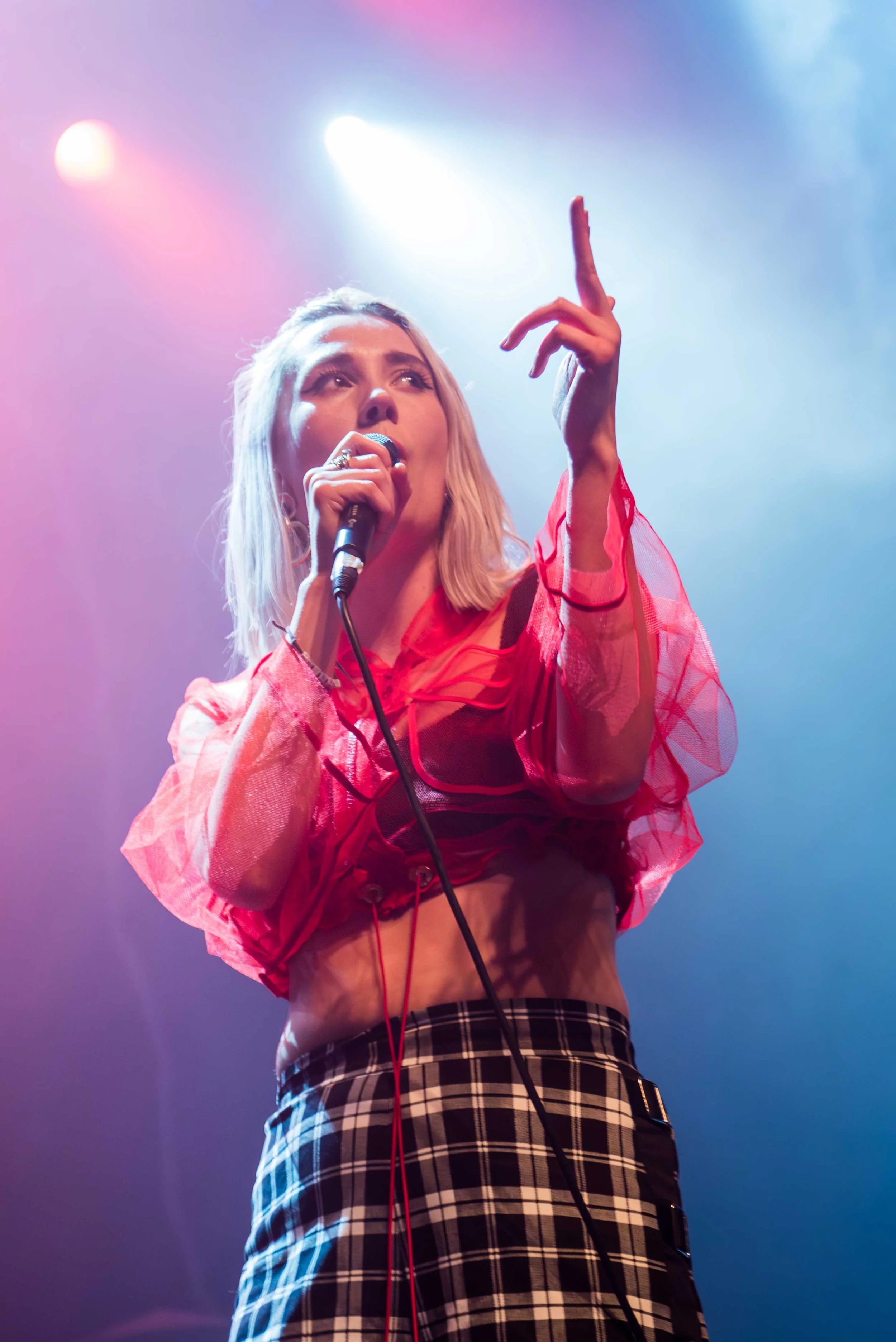 Yokana - Shot by Shotison. A female singer on stage holding a microphone, wearing a red see-through top with long sleeves and a black and white plaid skirt, with colourful stage lights overhead.
