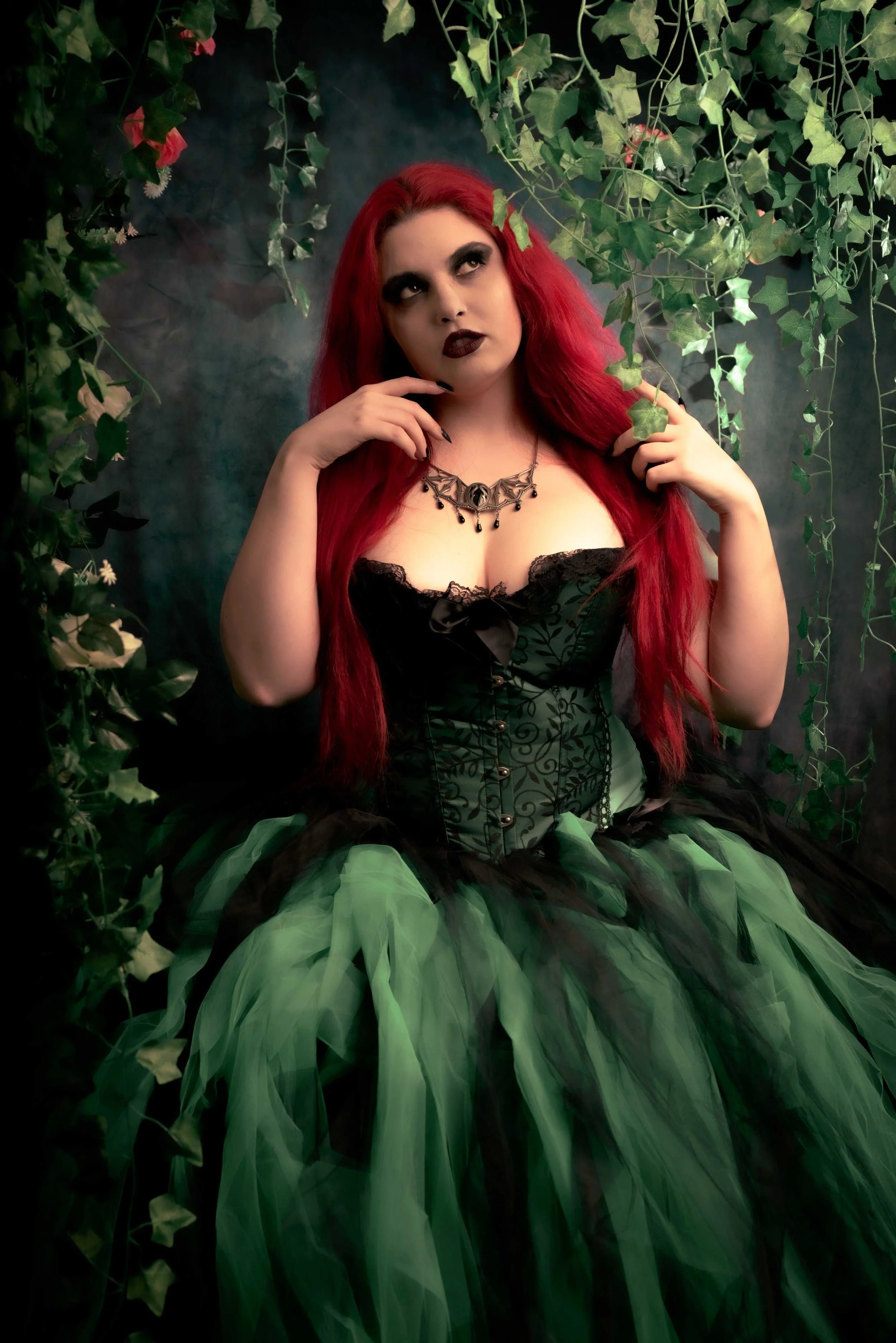 A woman with long red hair and dark makeup, wearing a gothic style black and green dress with lace and butterflies, posing among hanging green ivy leaves with a dark, smoky background.