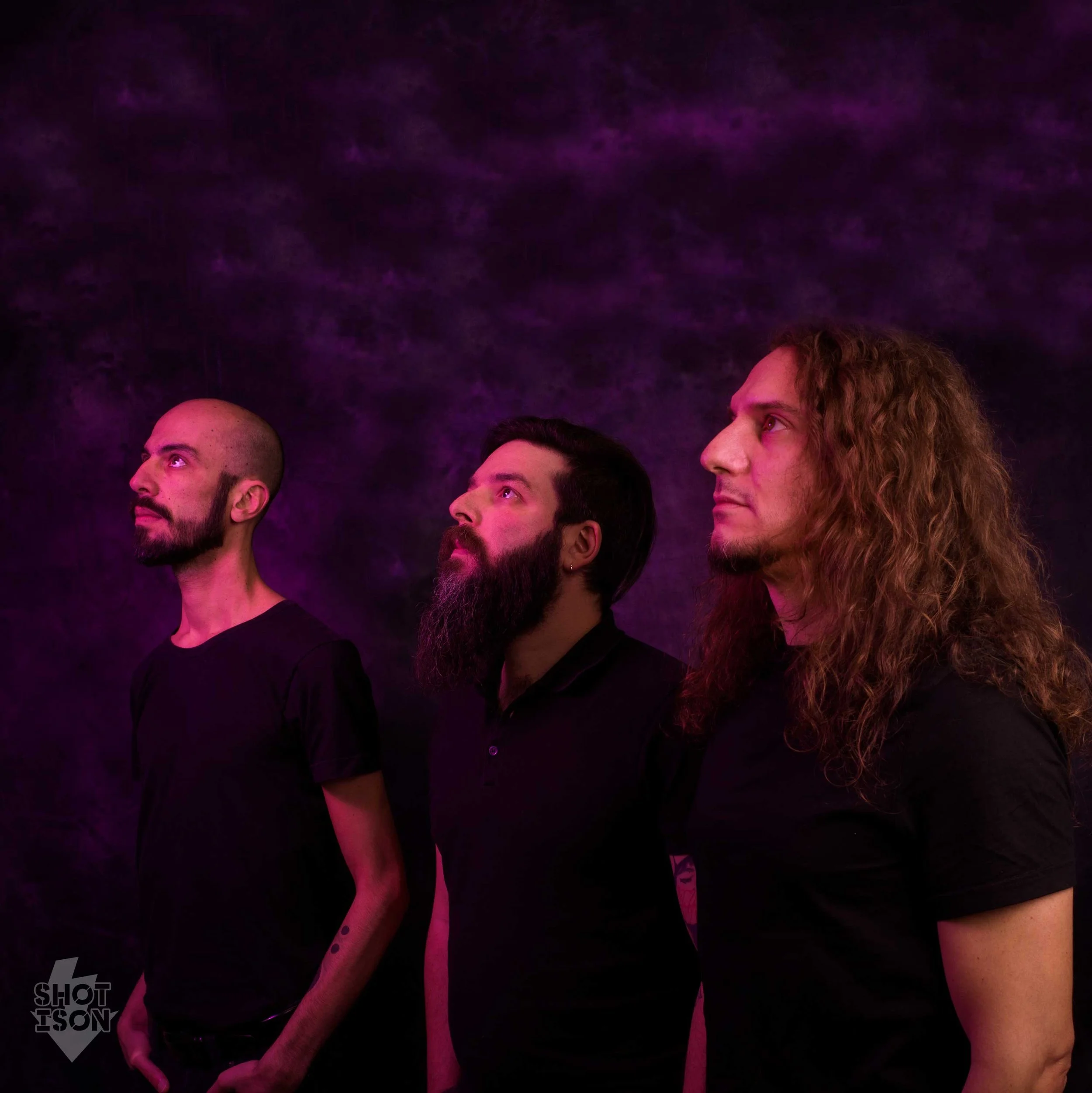 A group of people standing together in a dark setting with purple lighting, shown from the shoulders up, some looking to the side and others looking ahead.