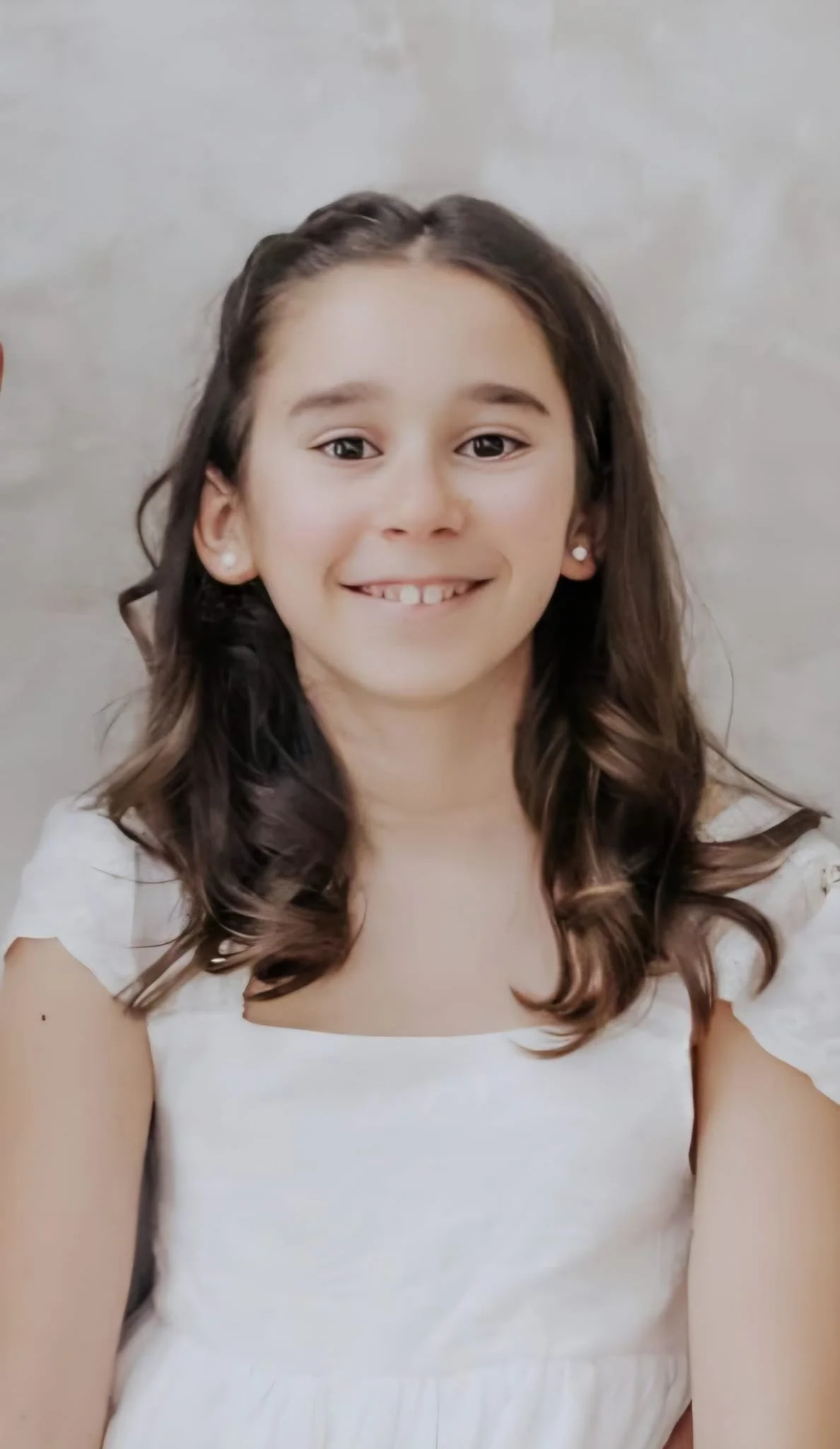 Retrato de una niña con cabello castaño oscuro y rizado, sonrisa y usando un vestido blanco, fondo gris claro.