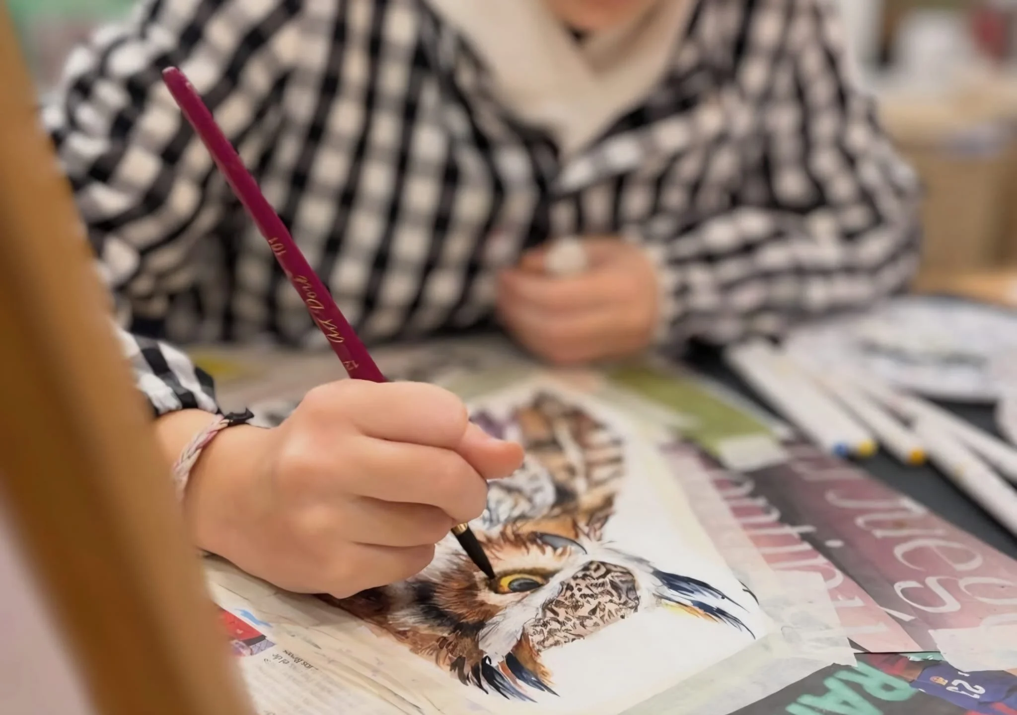 Manos sosteniendo un bolígrafo pintando un retrato de un ave, en una mesa con diferentes revistas y materiales de arte, en un entorno interior.
