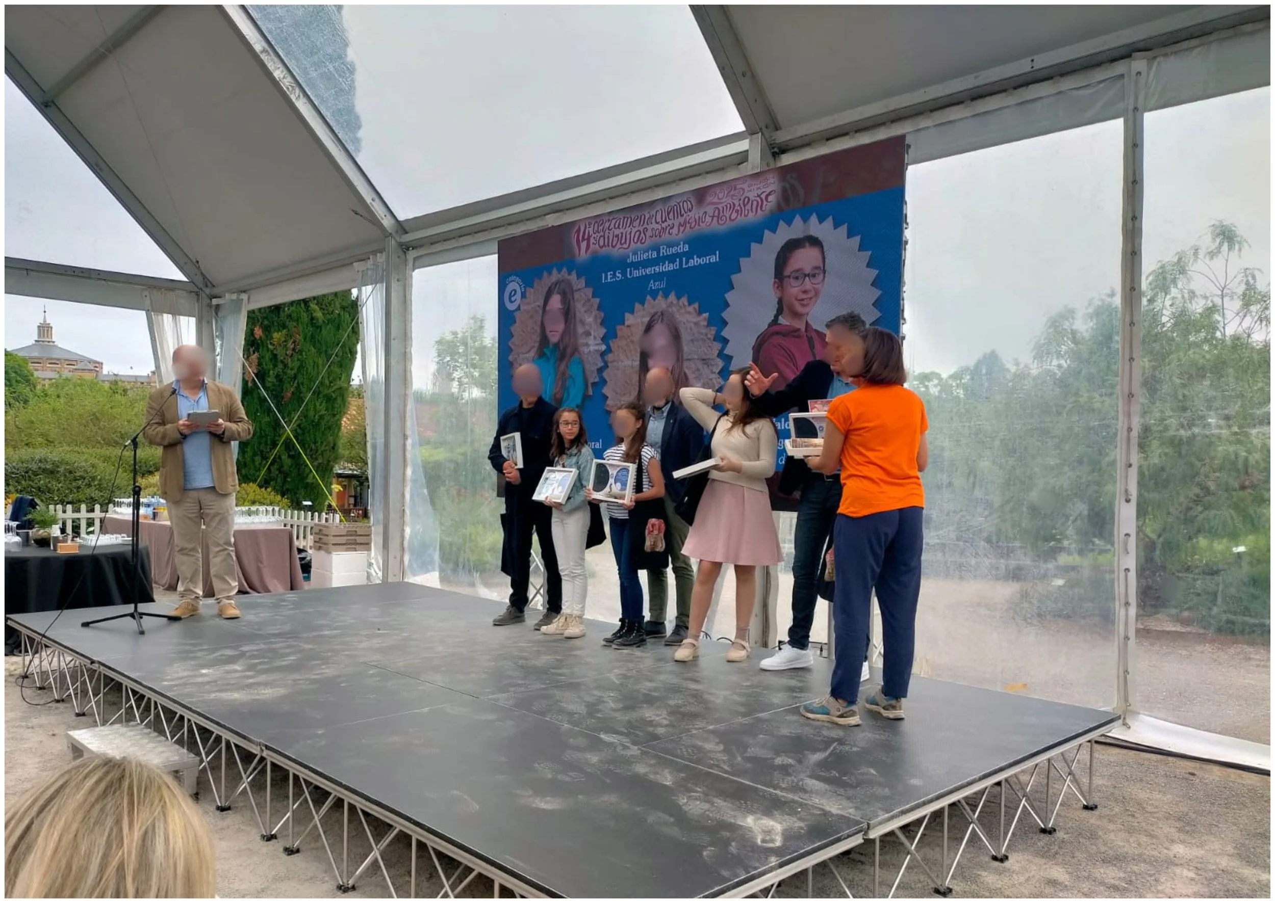 Personas en un escenario de celebración, con un cartel de fondo que muestra imágenes de niñas y texto, en un evento al aire libre, con presencia de una persona hablando y varias personas con libros o certificados.