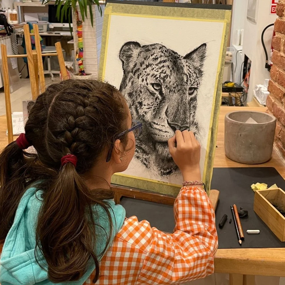 Una niña con trenza de cabello oscuro, usando una chaqueta azul y una camisa de cuadros naranja y blanco, está dibujando un leopardo en un caballete en un taller de arte.