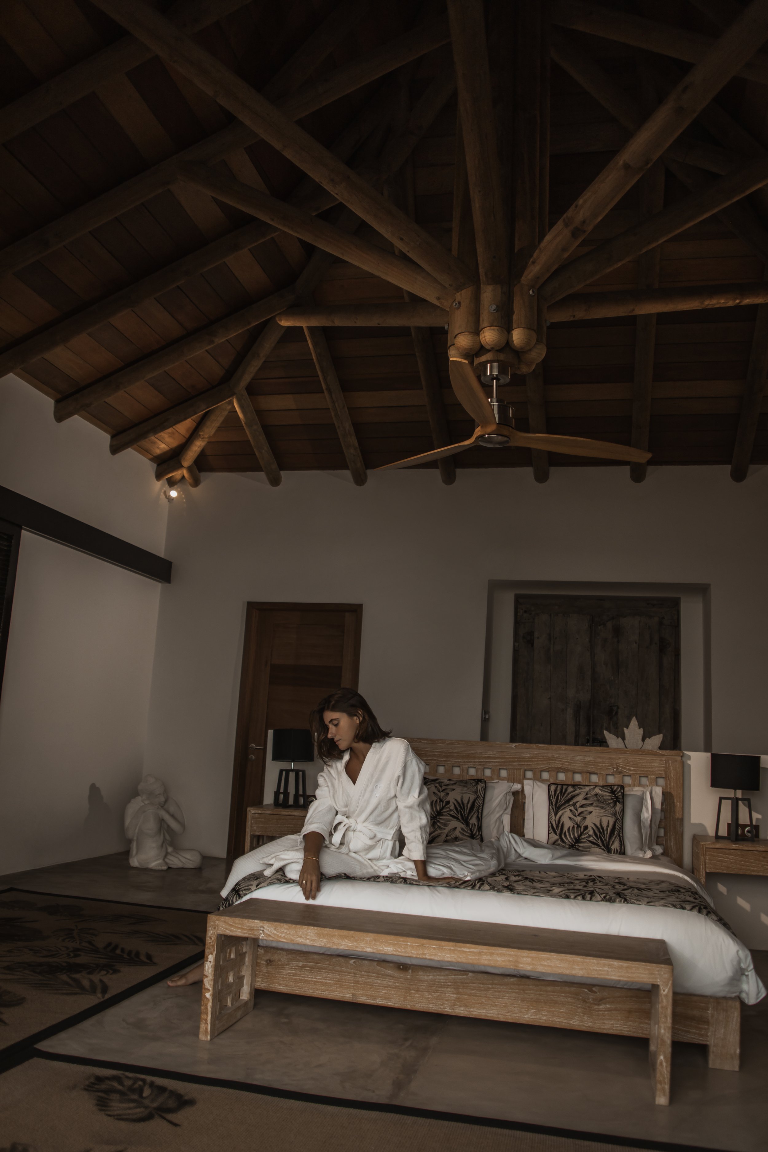 A woman in a white bathrobe sitting on a bed in a bedroom with wooden furniture and a wooden ceiling, holding her hand over her lap, with a Mona Lisa replica sculpture and a statue of a girl in the background.