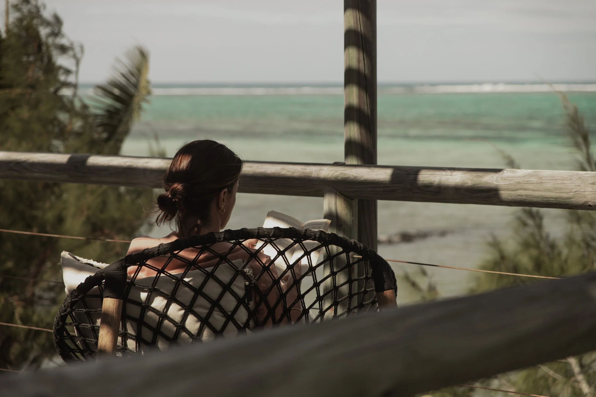 Woman with brown hair reading a book on a beachside deck with a view of the ocean and sky.