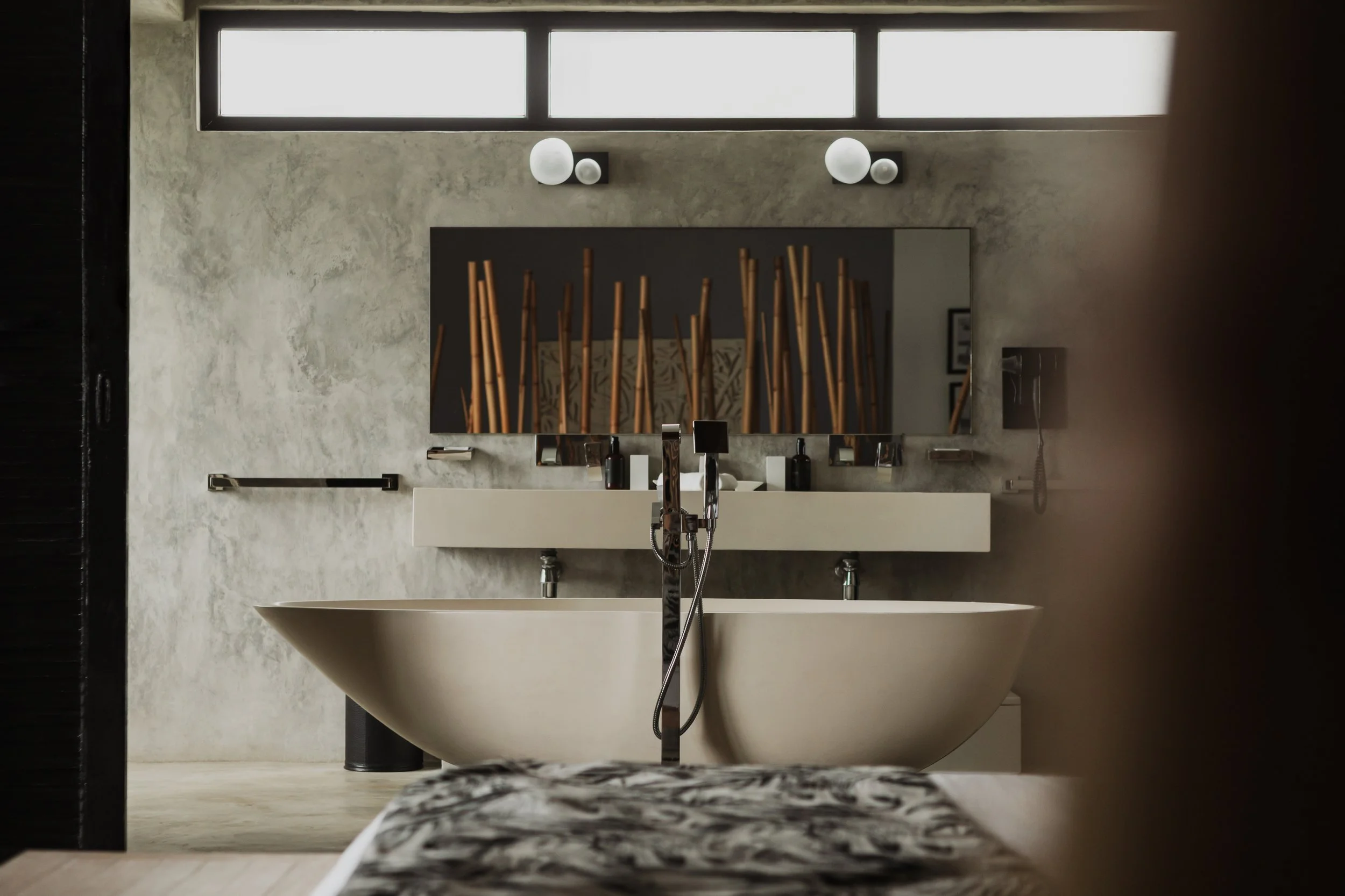Modern bathroom with a freestanding white bathtub, a large mirror, and a concrete wall. A window with frosted glass is above the mirror. There are black and white decorative items on the sink counter, and a wooden divider with sticks in the reflection of the mirror.