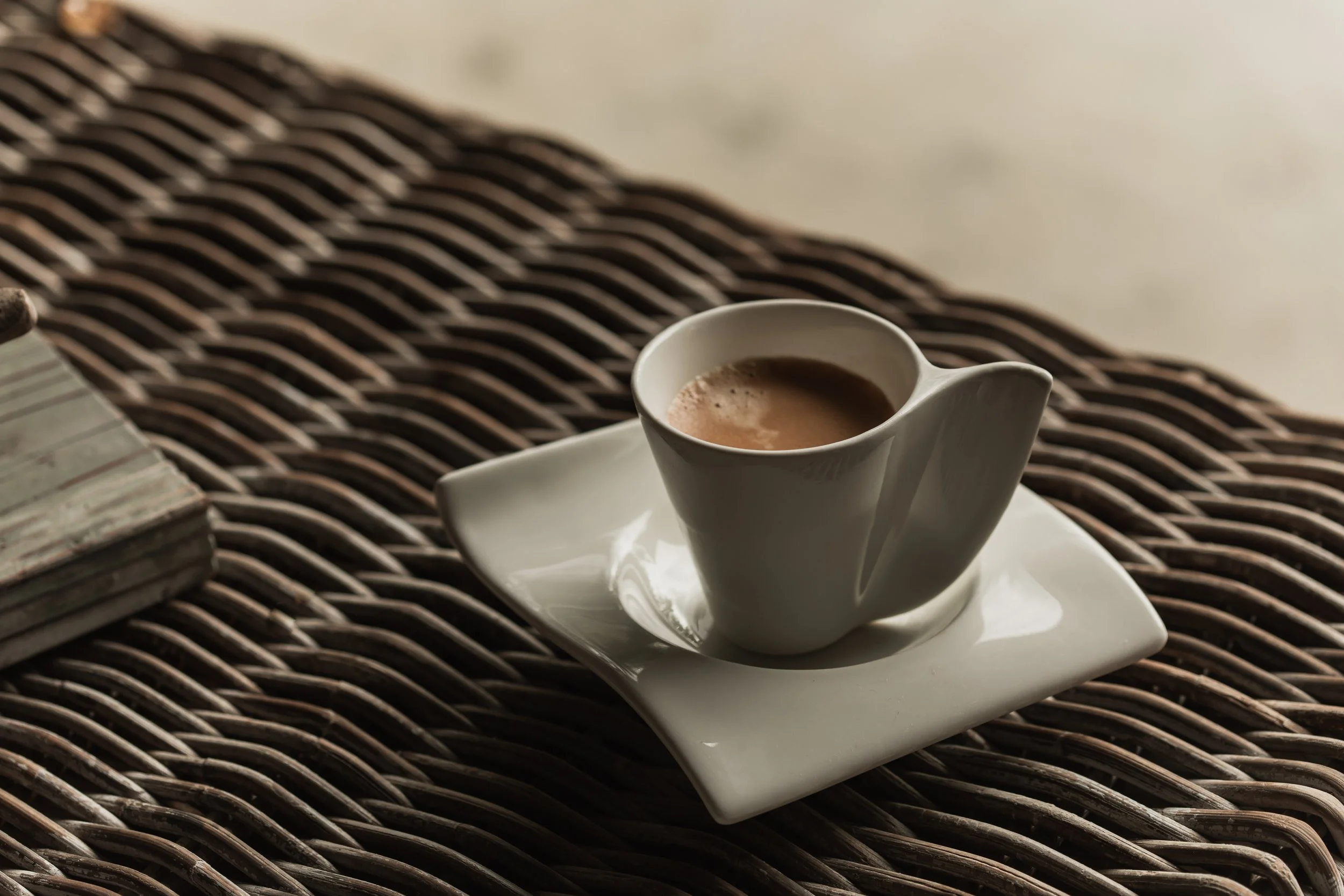A white porcelain cup of coffee with foam, placed on a white saucer with a wavy edge, on a dark woven surface with a book partially visible.