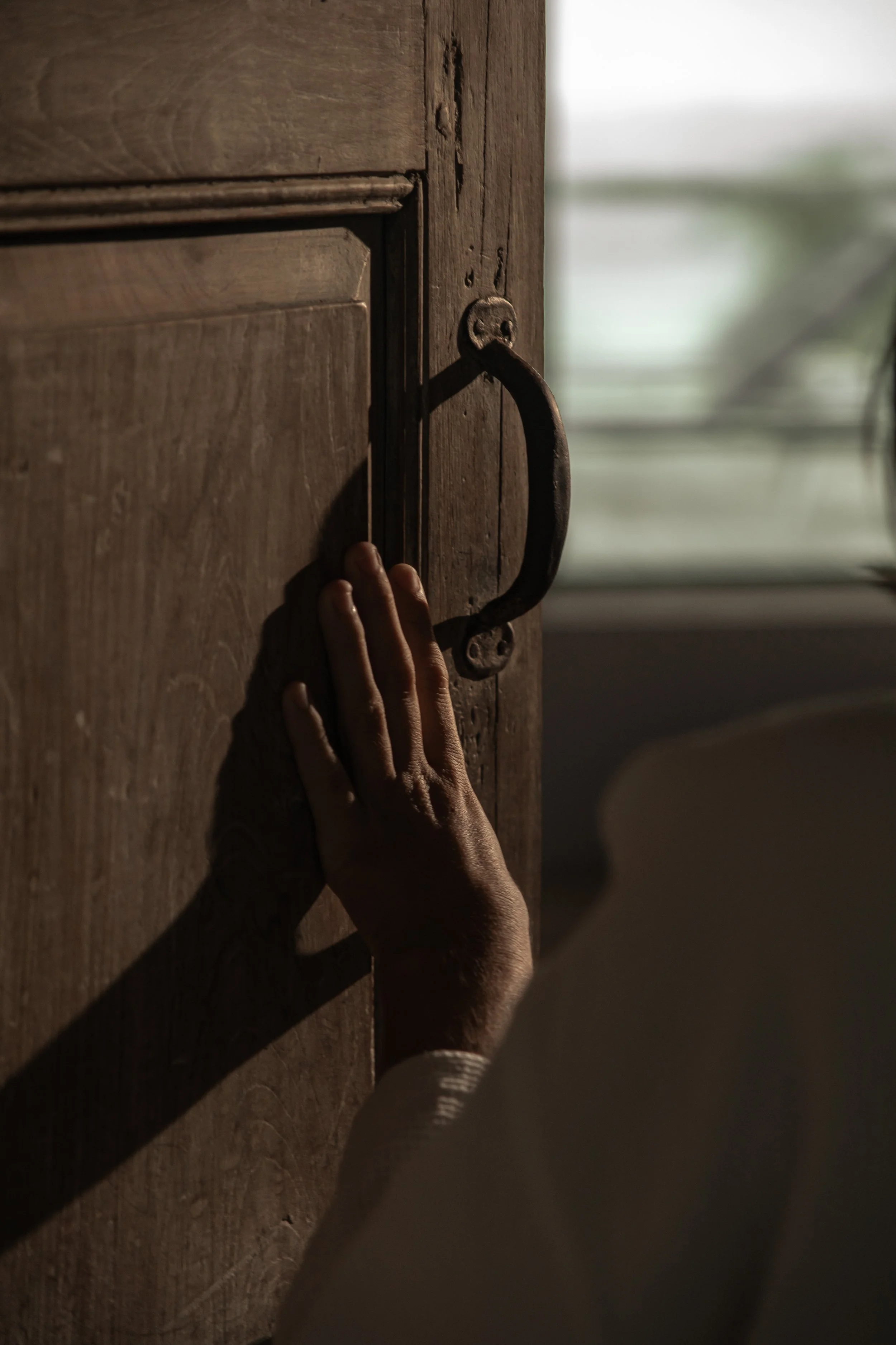 A person with a light-skinned hand touching a wooden door with a metal handle, with soft natural light coming through a window in the background.