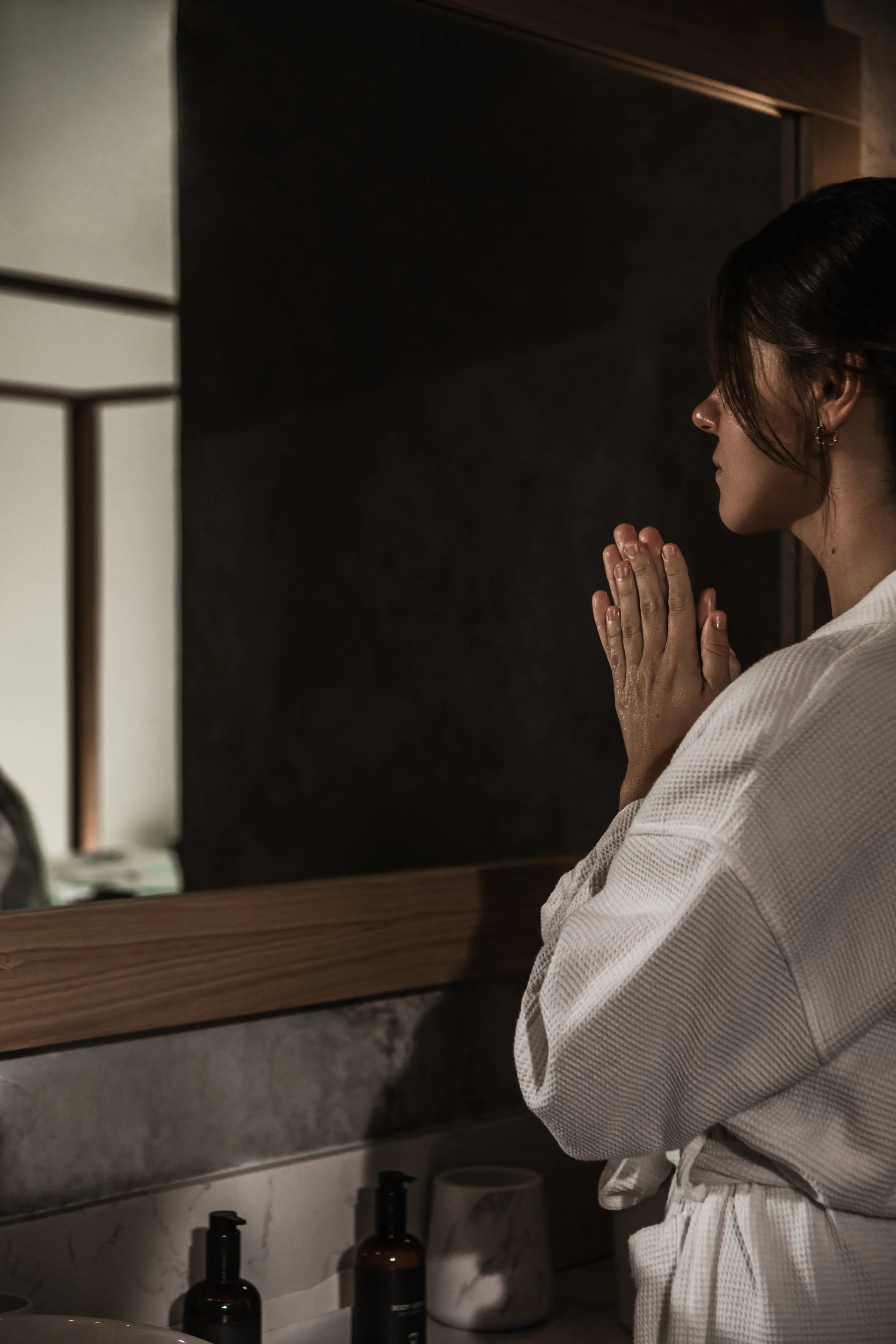 A woman with dark hair tied back, wearing a white bathrobe, stands in front of a bathroom mirror with her hands pressed together in prayer or meditation. The bathroom has a marble countertop with two black bottles, a white electric appliance, and a beige container. The woman is gazing at her reflection with a calm expression.