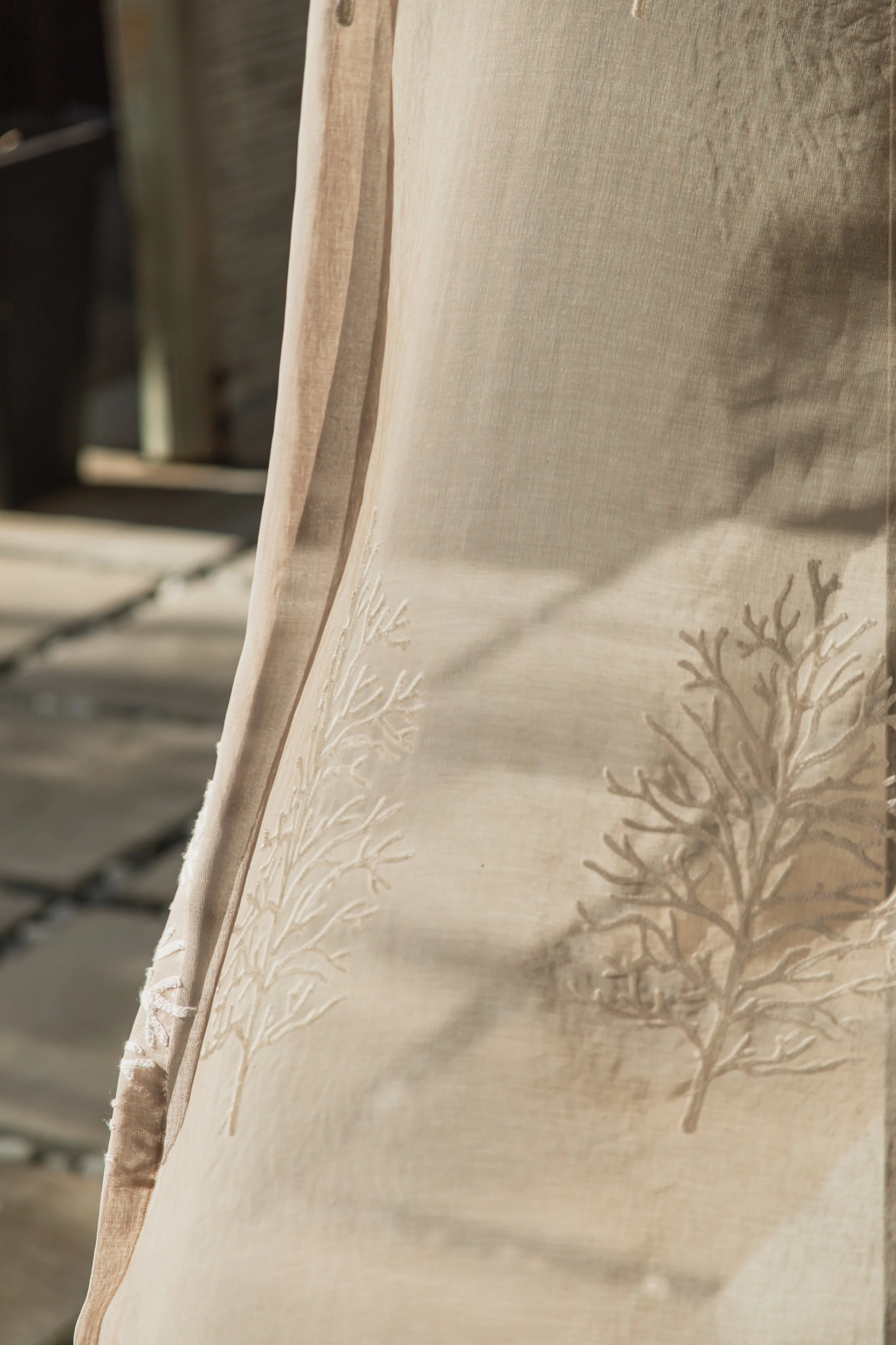 Close-up of a light-colored fabric curtain with a subtle embroidered tree branch pattern, hanging vertically.