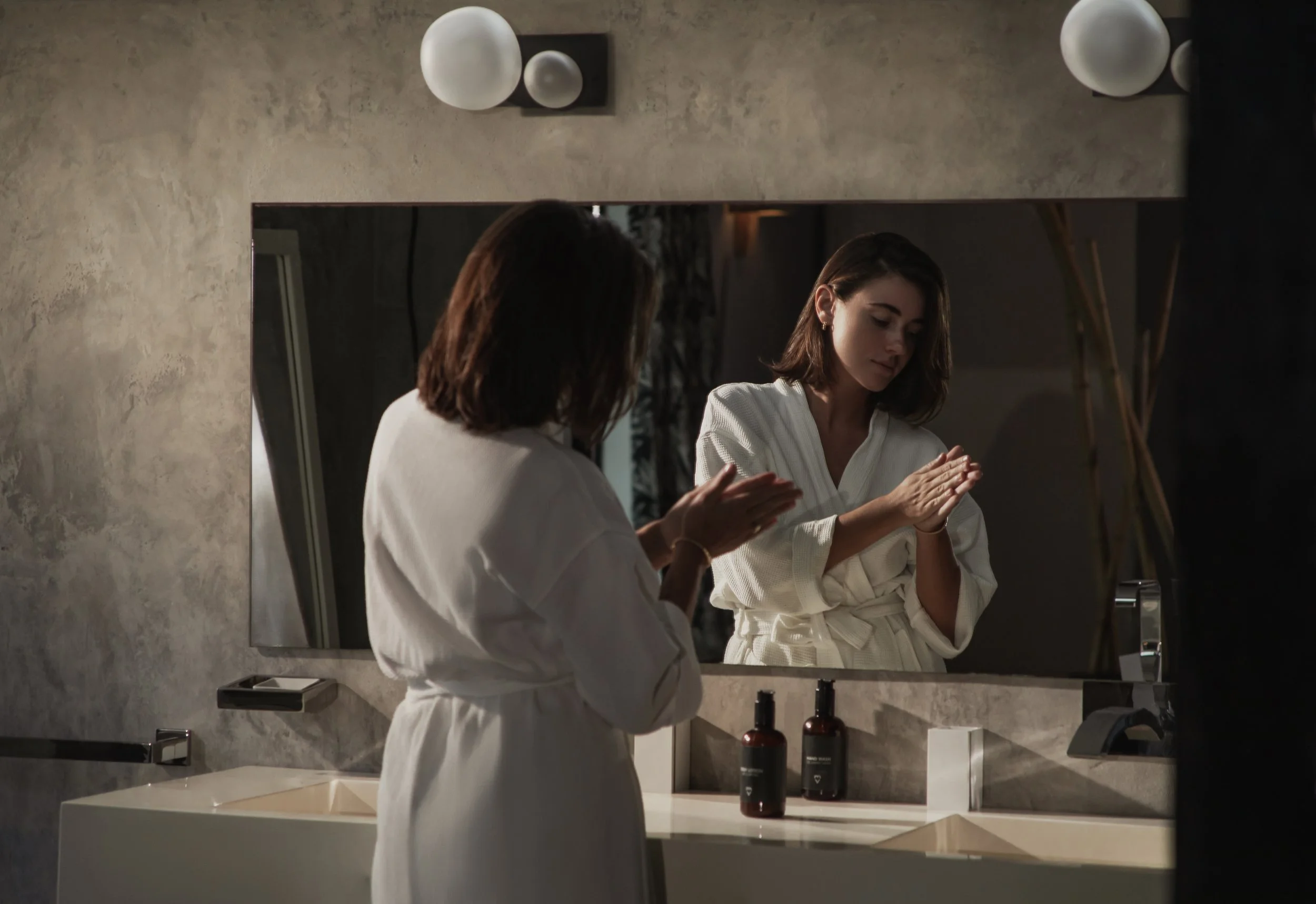 A woman in a white bathrobe looks at her reflection in a bathroom mirror while adjusting her sleeves.