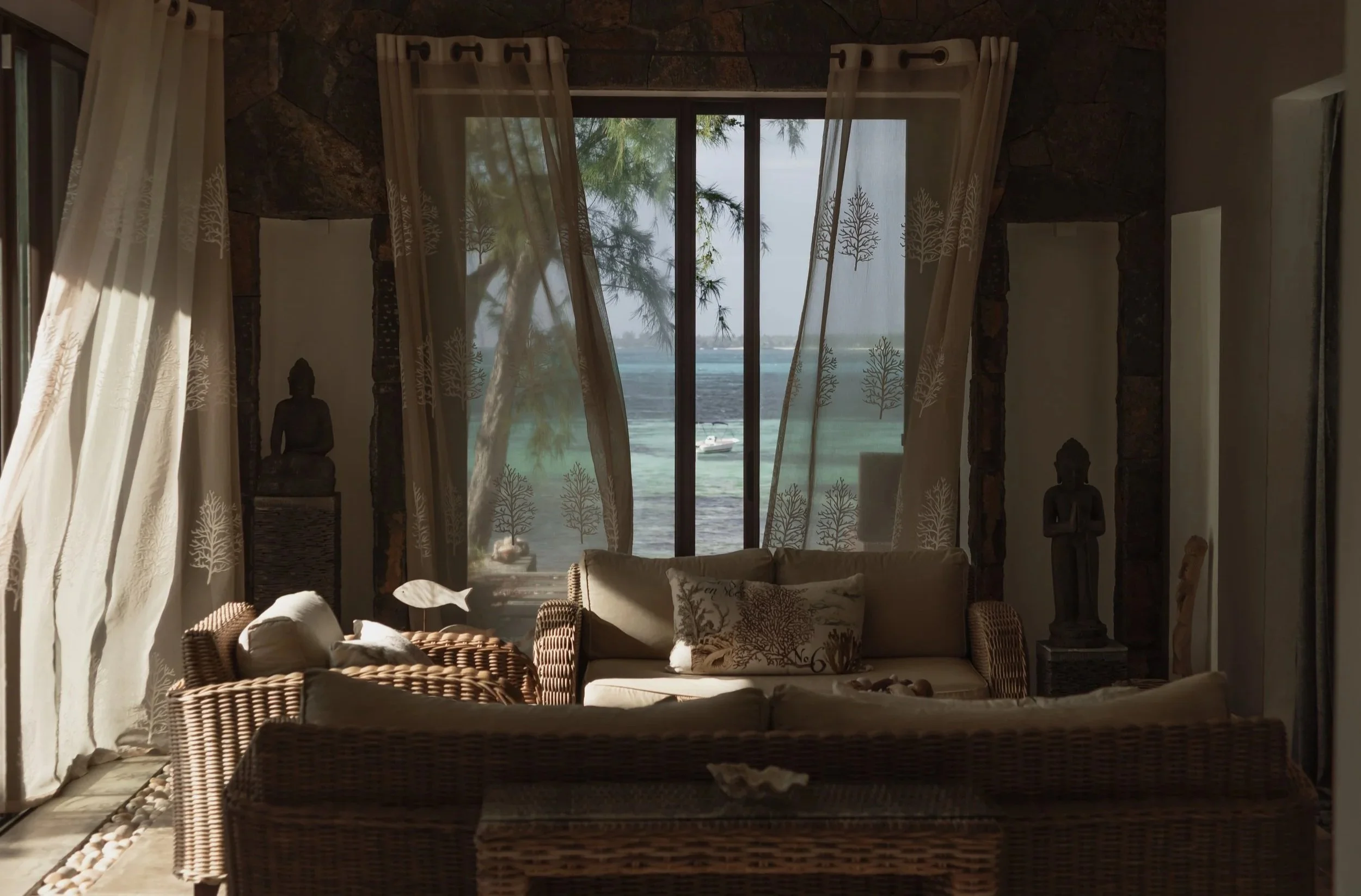 Interior of a room with wicker furniture, curtains, and sculptural decor, overlooking a body of water with a boat and trees outside.