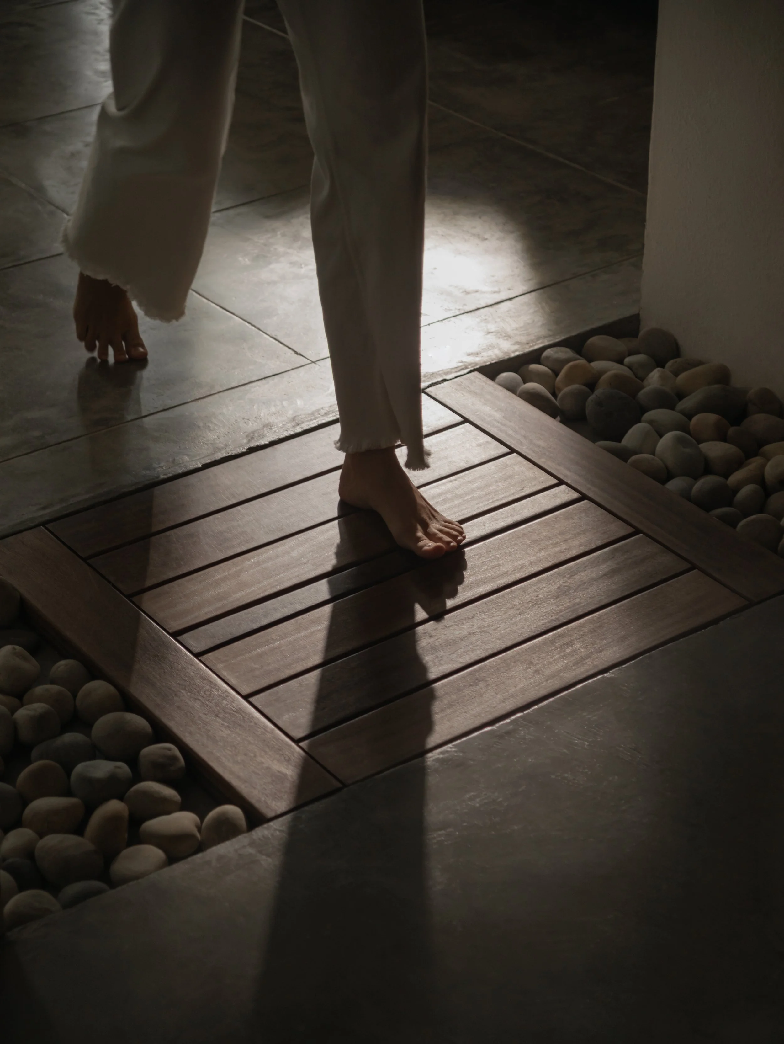 Partial view of a person walking barefoot on a tiled floor, with visible white pants and a long sleeve shirt, near a small section of white wall with a collection of round stones.