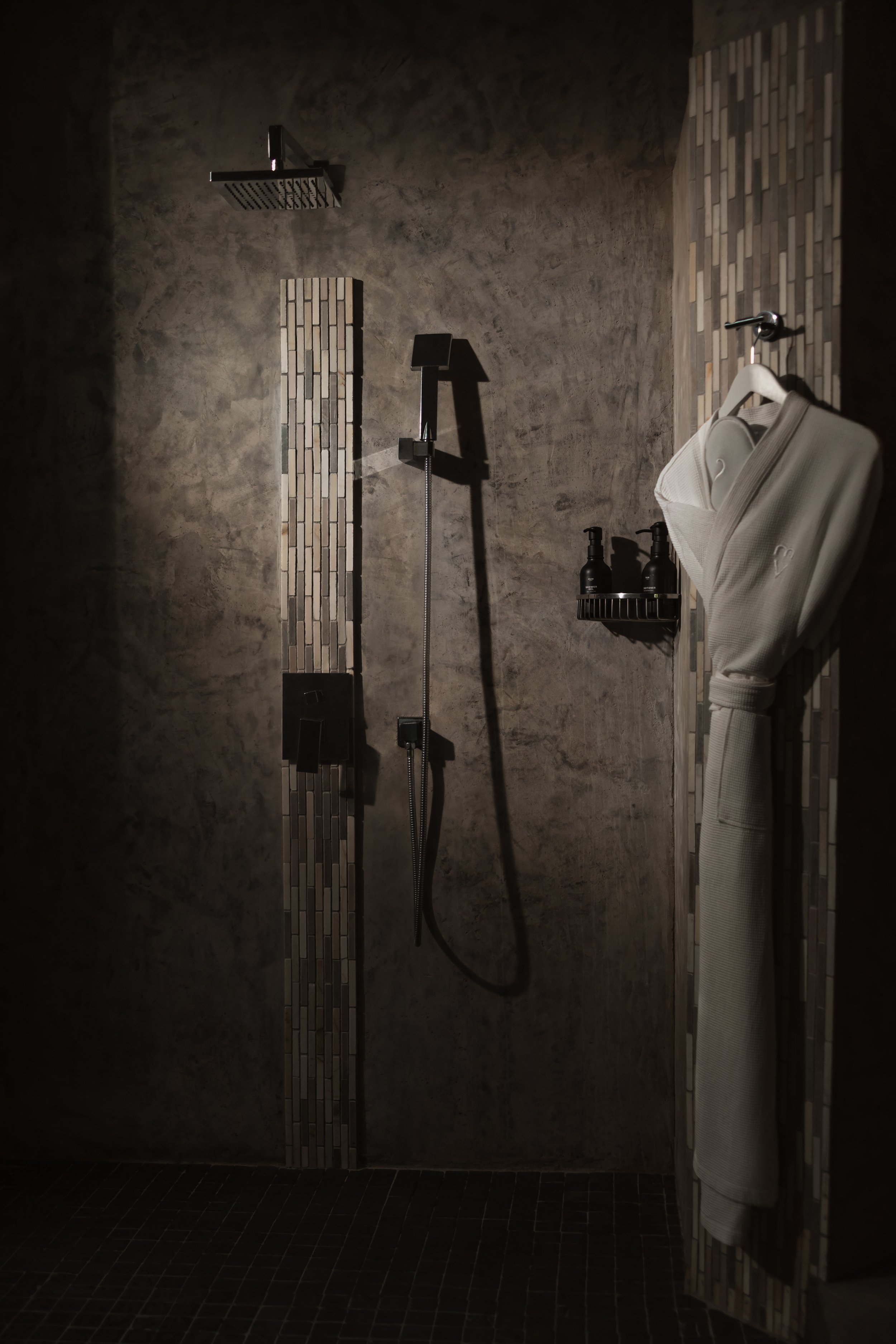 A modern shower area with dark tiled walls and floor. The showerhead is mounted on the ceiling. There is a sleek, black control panel on the wall and two bottles of shower products on a small shelf. A bathrobe hangs on a hook to the right.