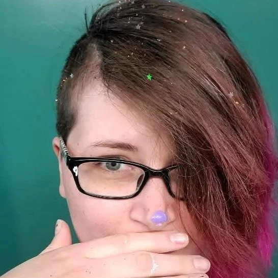 A person with short hair and glasses partially covering their face, with confetti glitter and pink hair dye, against a teal background.