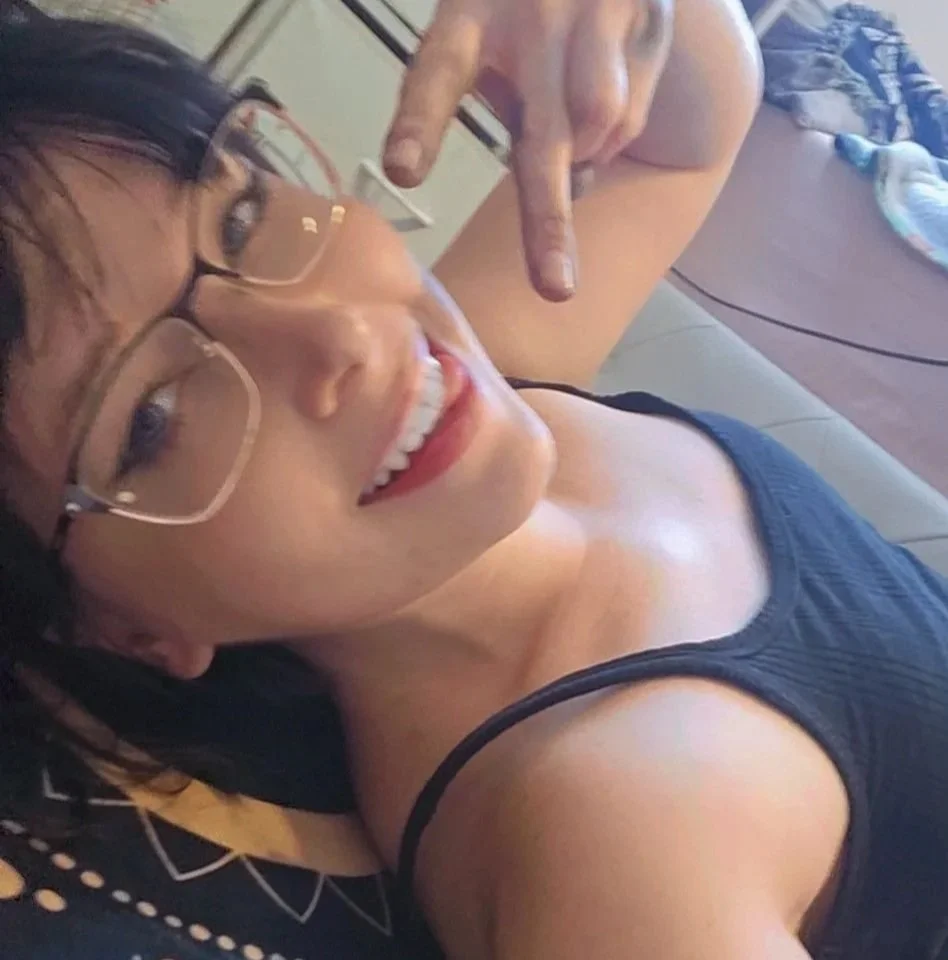 A woman with glasses lying on the floor, smiling, and holding up 2 fingers.
