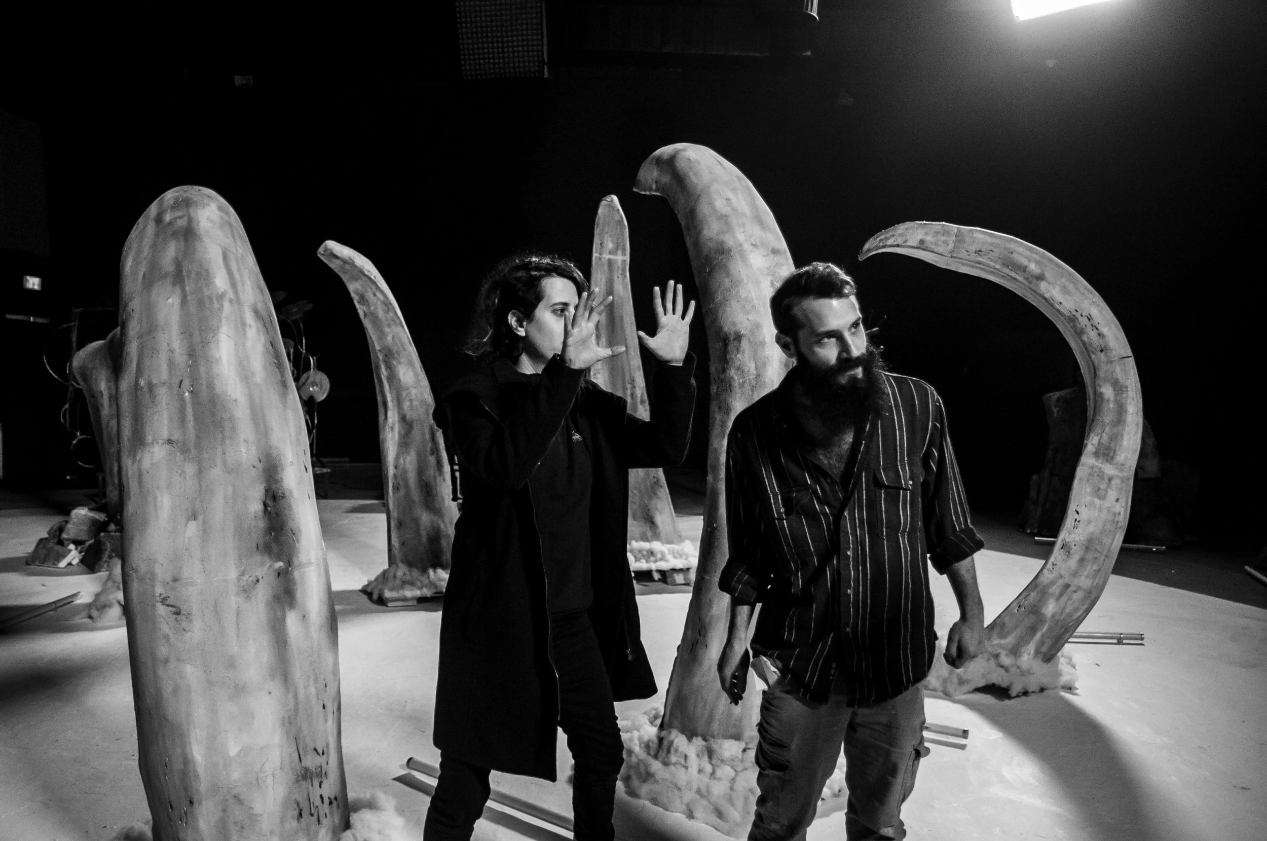 Two people, a woman with short hair and a beard man, stand among large bamboo or wood structures in a dark, indoor space. The woman is gesturing with her hands while talking, and the man is looking downward, music video director , Music video clip