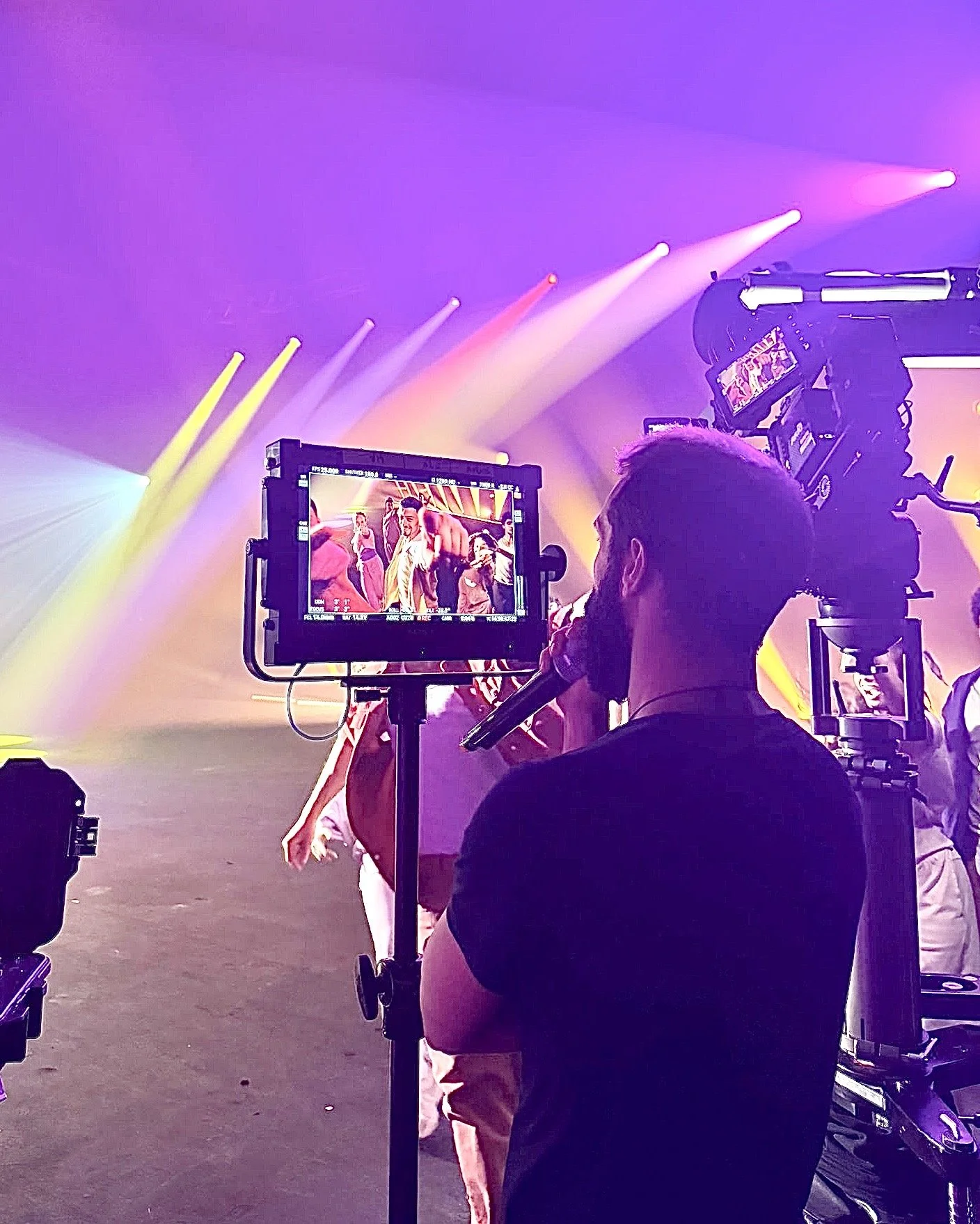 Behind-the-scenes view of a video shoot, showing a camera operator filming a performer on stage with colorful stage lights and smoke effects. Avi Mazliah on set BTS
