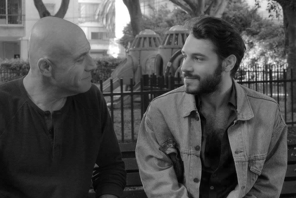 Two men sitting on a park bench, facing each other and smiling. Background shows playground equipment and trees. heritage movie by Yuval Aharoni