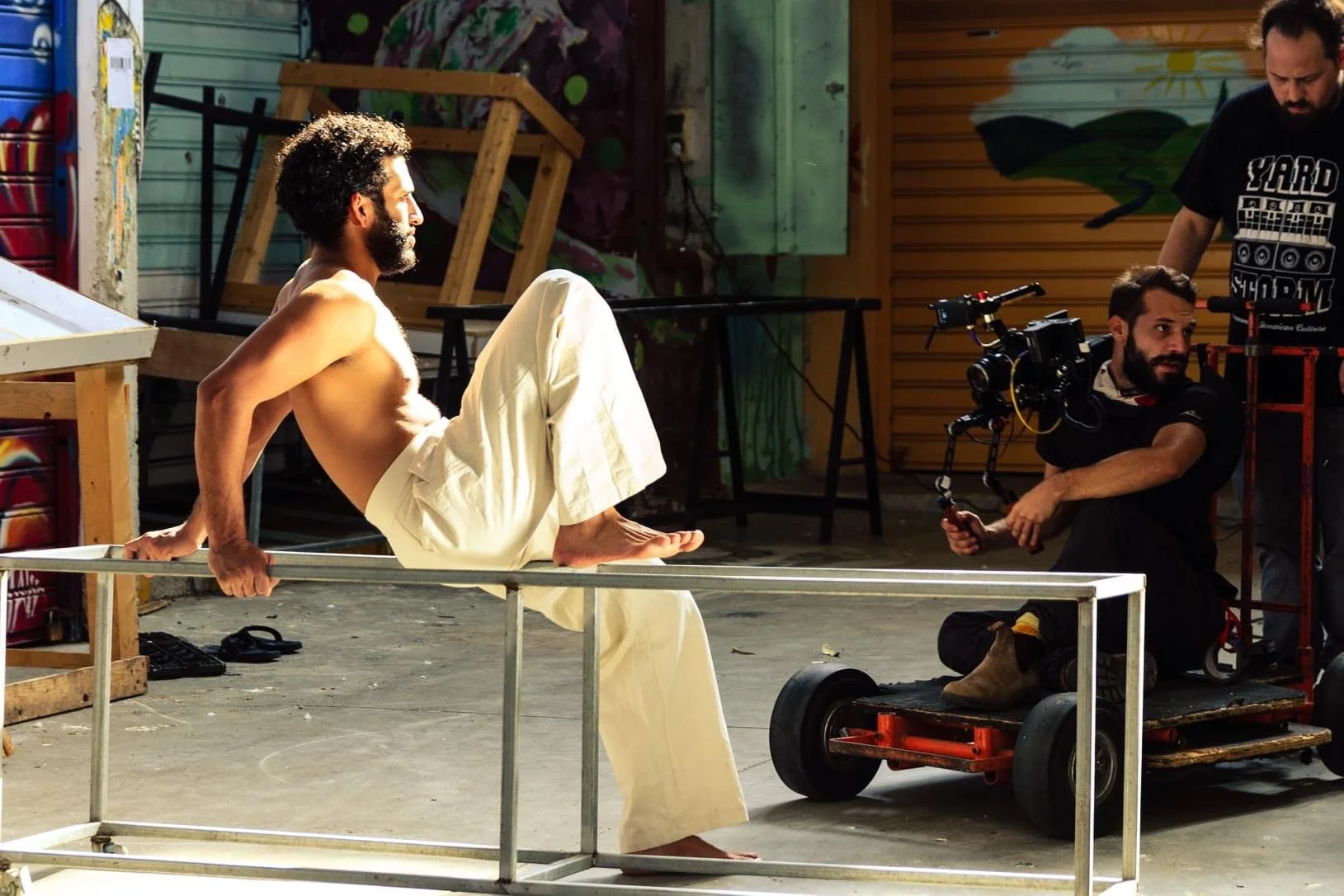 A shirtless man with a beard and curly hair is sitting on a metallic frame, in a casual setting with graffiti on the wall behind him.. Bar Tzabari Music Video HADERA, Hadera, Behind the seance, music video director 