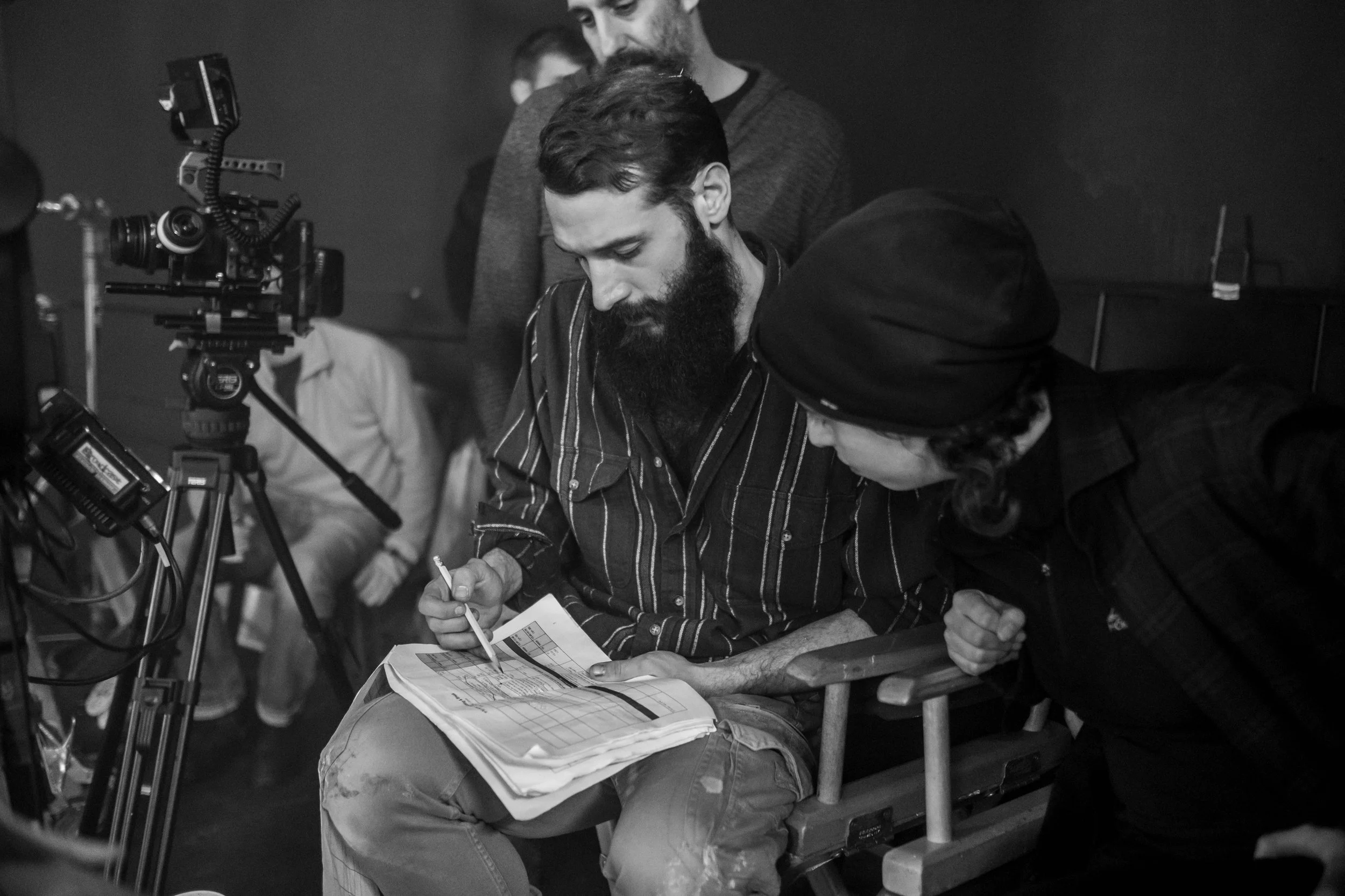 Black and white photo of a group of people in a professional setting. A man with a beard, striped shirt, and gloves is sitting on a bench, focused on writing or drawing in a notebook placed on his lap. music video director , Music video clip