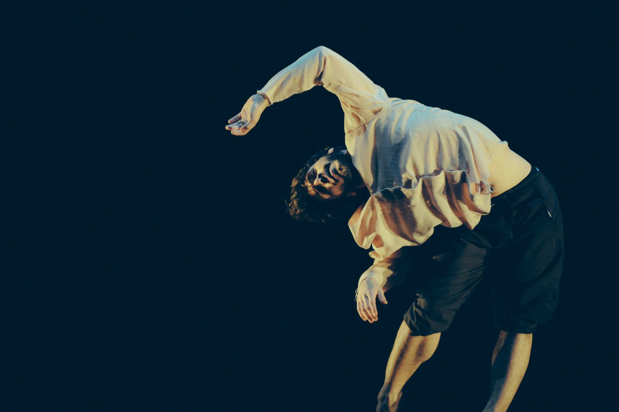 A person with curly hair wearing a loose, long-sleeved shirt and dark pants, performing a dance move on a dark stage, with their body bent backward and arms extended.  Avi Mazliah dance dancing performance 