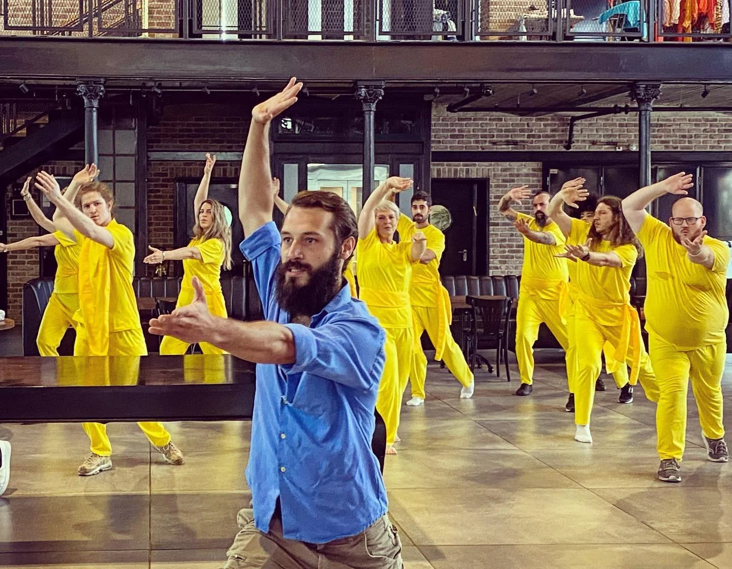 A group of people dressed in yellow matching outfits practicing martial arts or dance moves under a trainer in a gym or studio with brick walls and large mirrors.