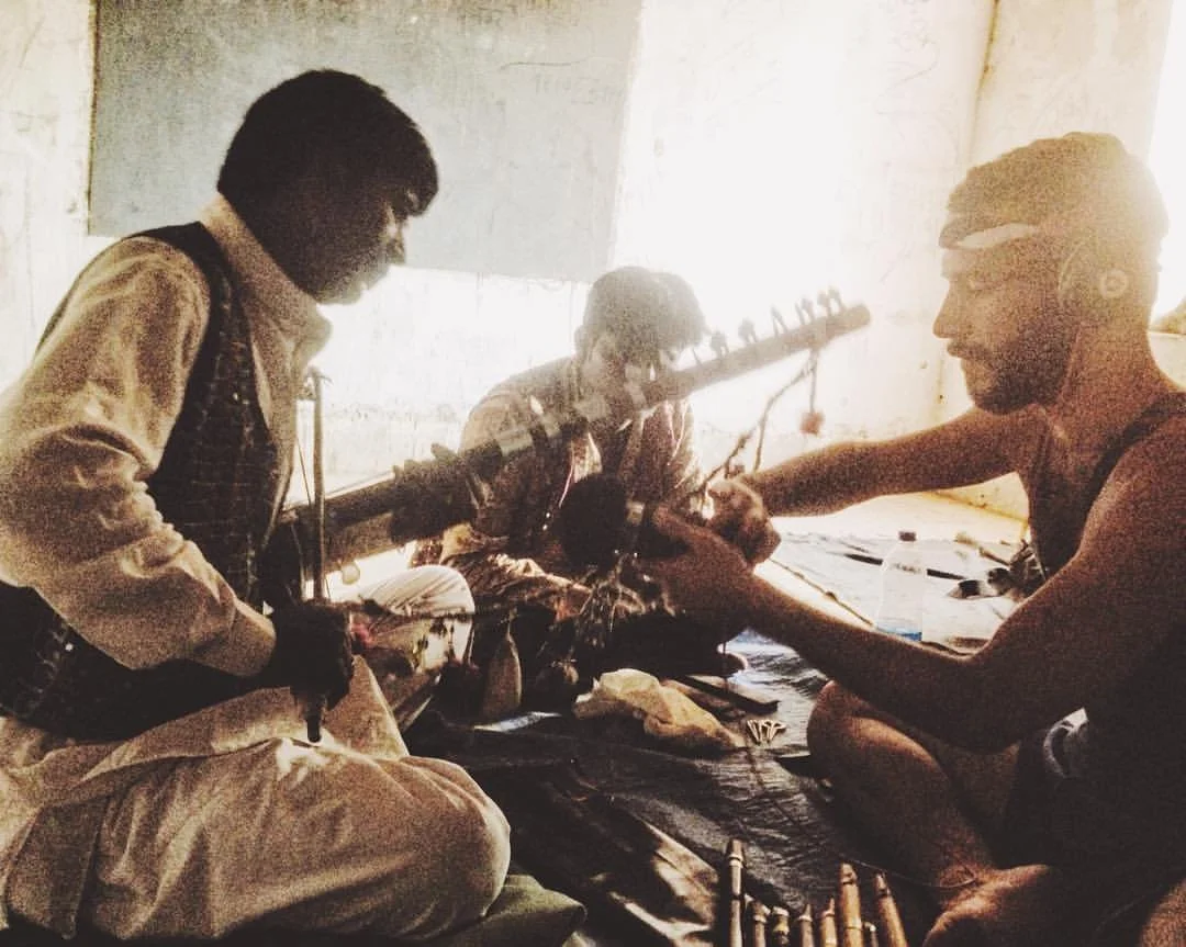 Three men are sitting on the floor during a music session, with two playing guitars and one assisting with equipment in a simple room illuminated by natural light.