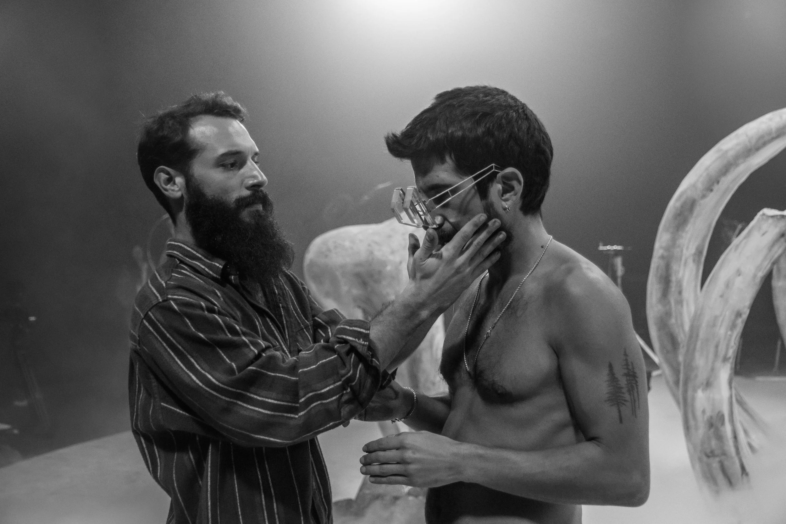 Two men are in a scene resembling a tattoo or body art studio. One man, with a beard and striped shirt, is applying a tattoo or piercing to the other man's chest, music video director , Music video clip