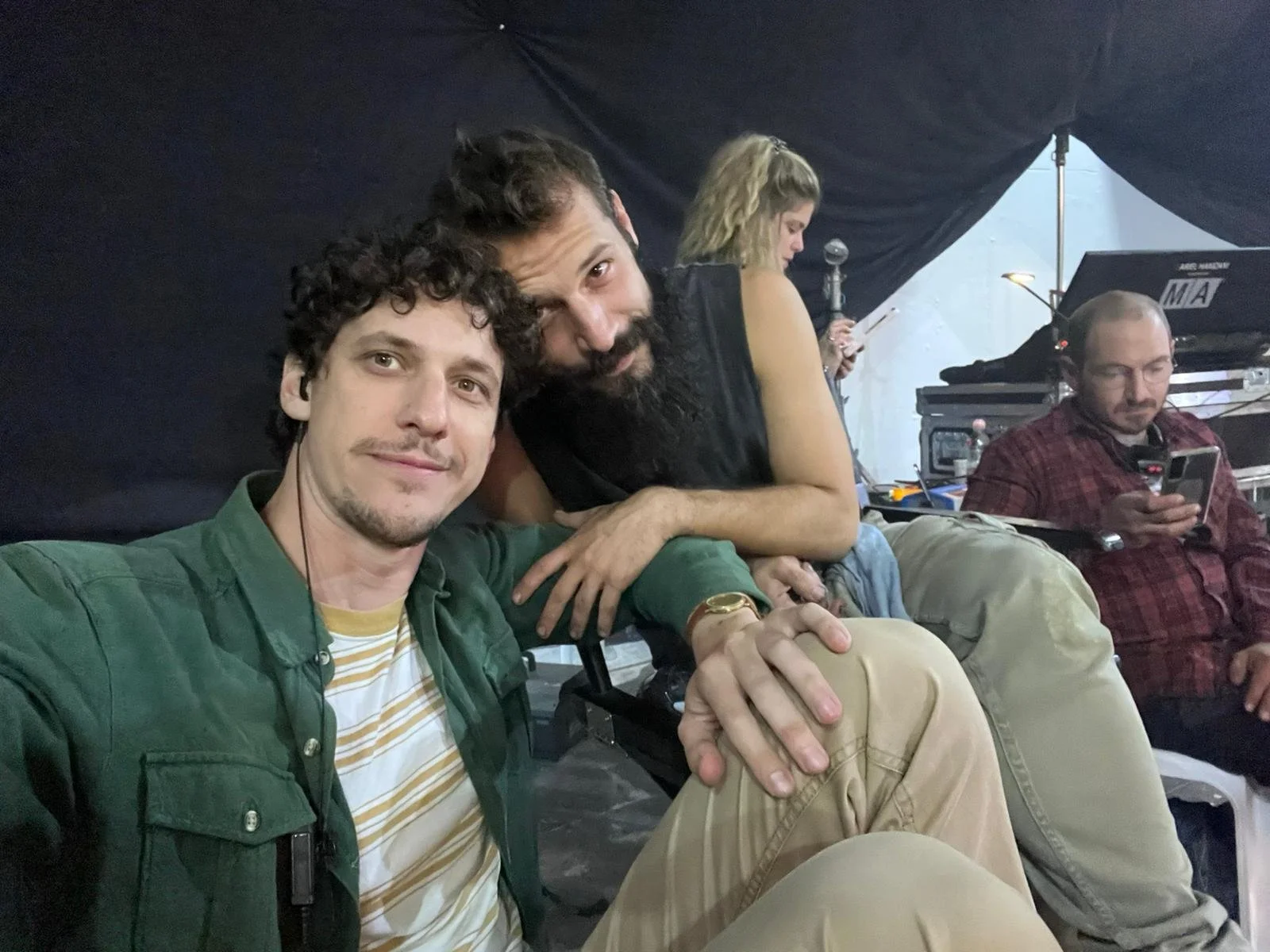 Group of three people sitting in a studio or backstage area, with equipment visible in the background. Two men are in the foreground, one with curly hair and the other with a beard, both smiling slightly. A woman in the background is using her phone,
