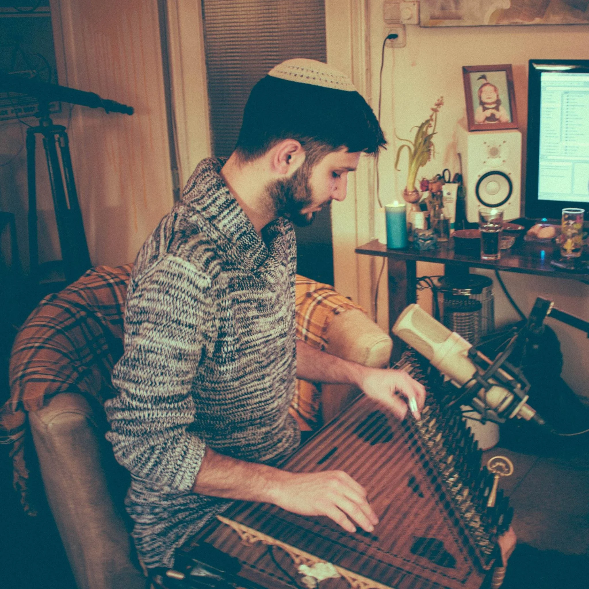 A man sitting at a wooden dining table in a cozy room, playing Kanun wearing a patterned sweater and a kippah, with drinks and snacks on the table, and musical equipment visible in the background.