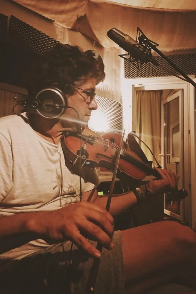 A person in a recording studio, playing the violin and wearing headphones, with a microphone overhead.