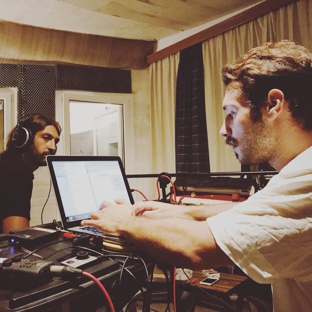 Two men in a recording studio. One man with headphones sitting at a table with audio equipment, and another man using a laptop. The room has soundproofing and curtains.