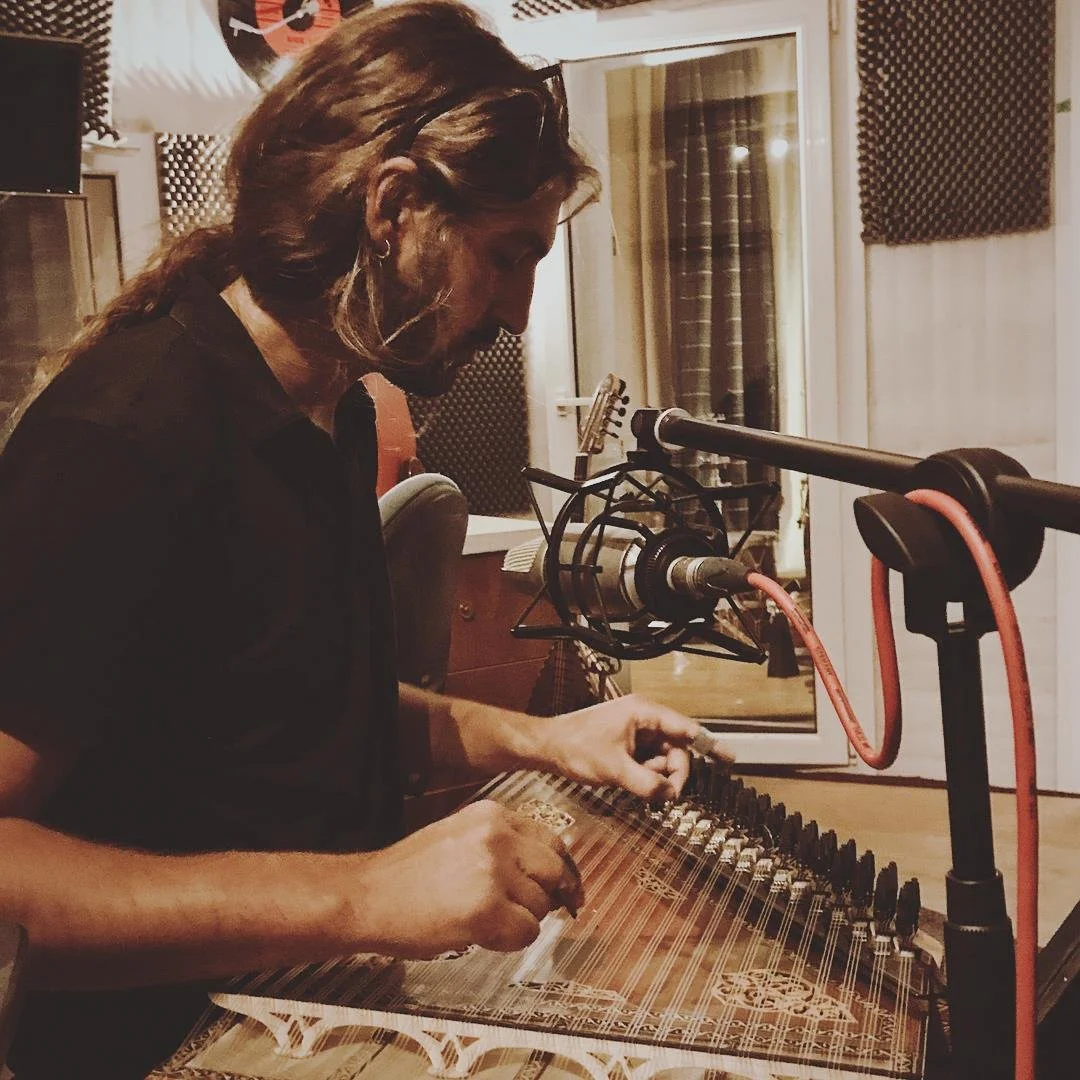 Man with long hair and earrings working in a recording studio, wearing a black shirt, using a metal guitar, Kanun , microphone in front, surrounded by soundproofing foam.