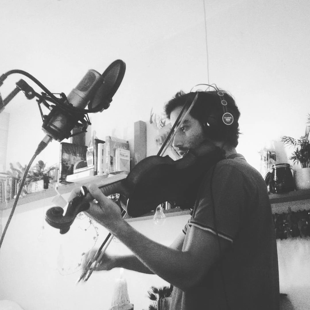 A young man wearing headphones playing a violin in a room with bookshelves and decorated with plants. recording album