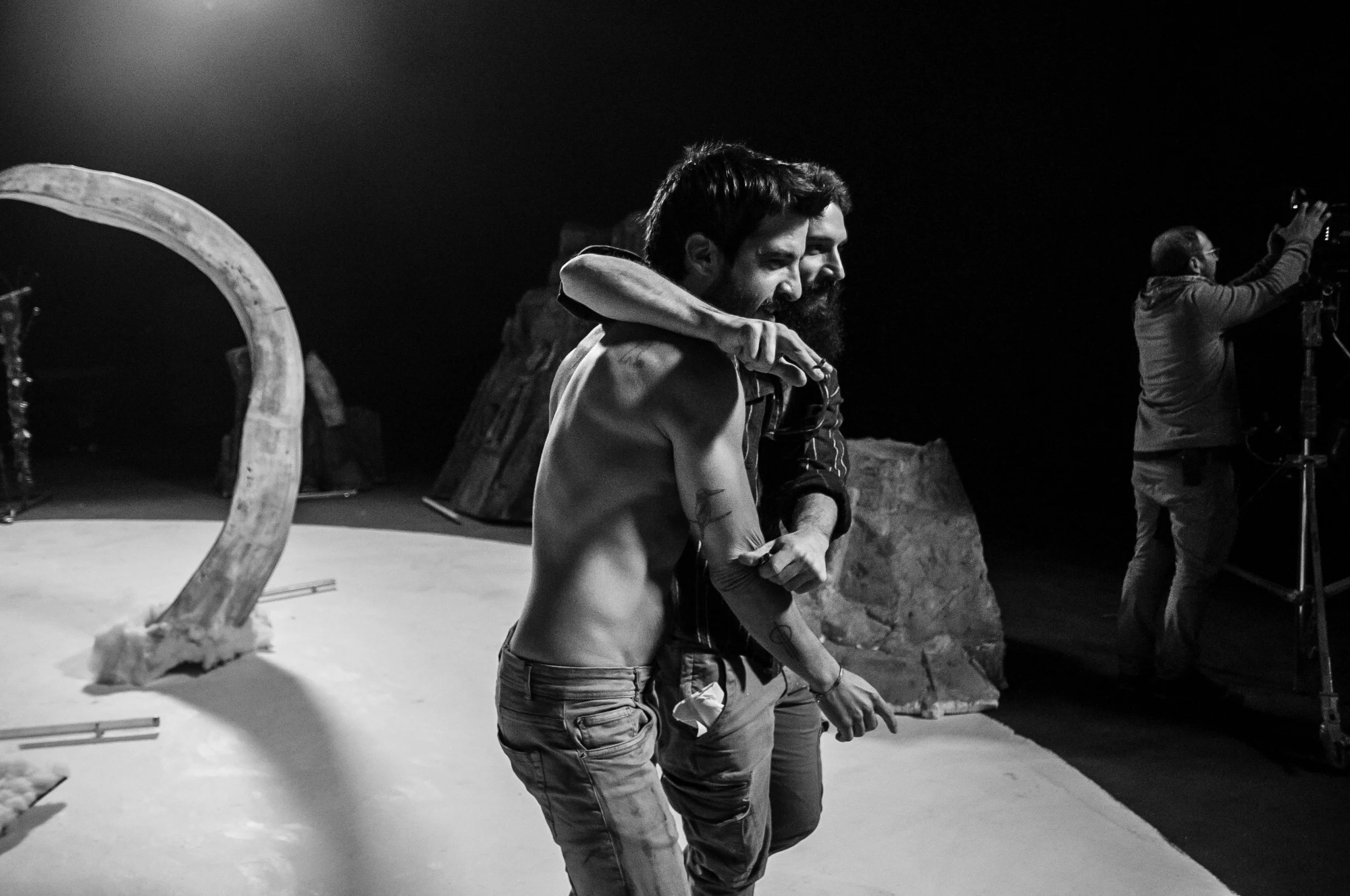 Two men embracing, smiling, in a room with a dark background. One man has a beard and tattoos, the other has a beard and dark hair. Another person with glasses is taking a photo in the background. music video director , Music video clip