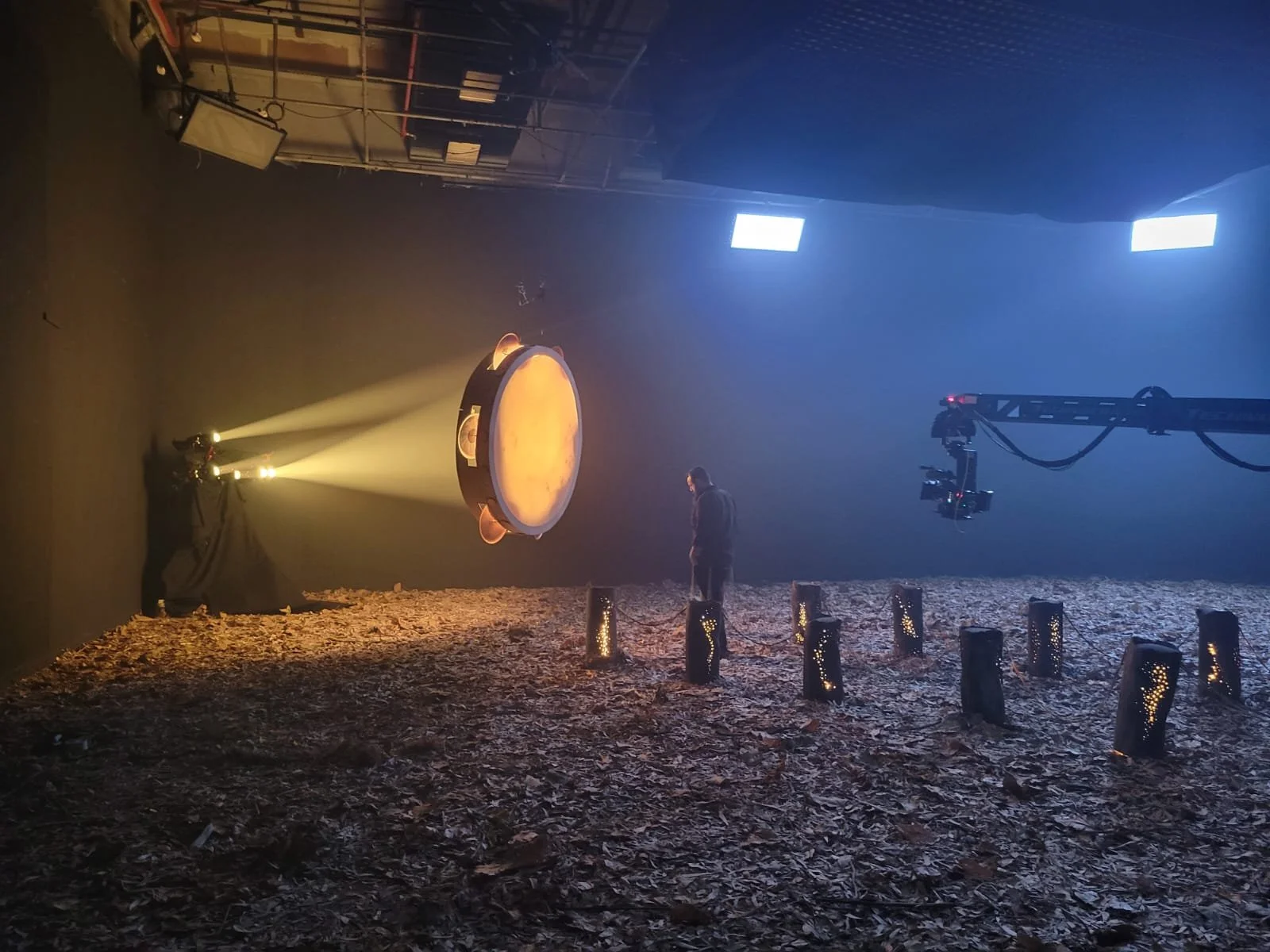Set of stage lights shining on a person and a large circular object suspended in a dark room with a textured floor and a camera crane visible.