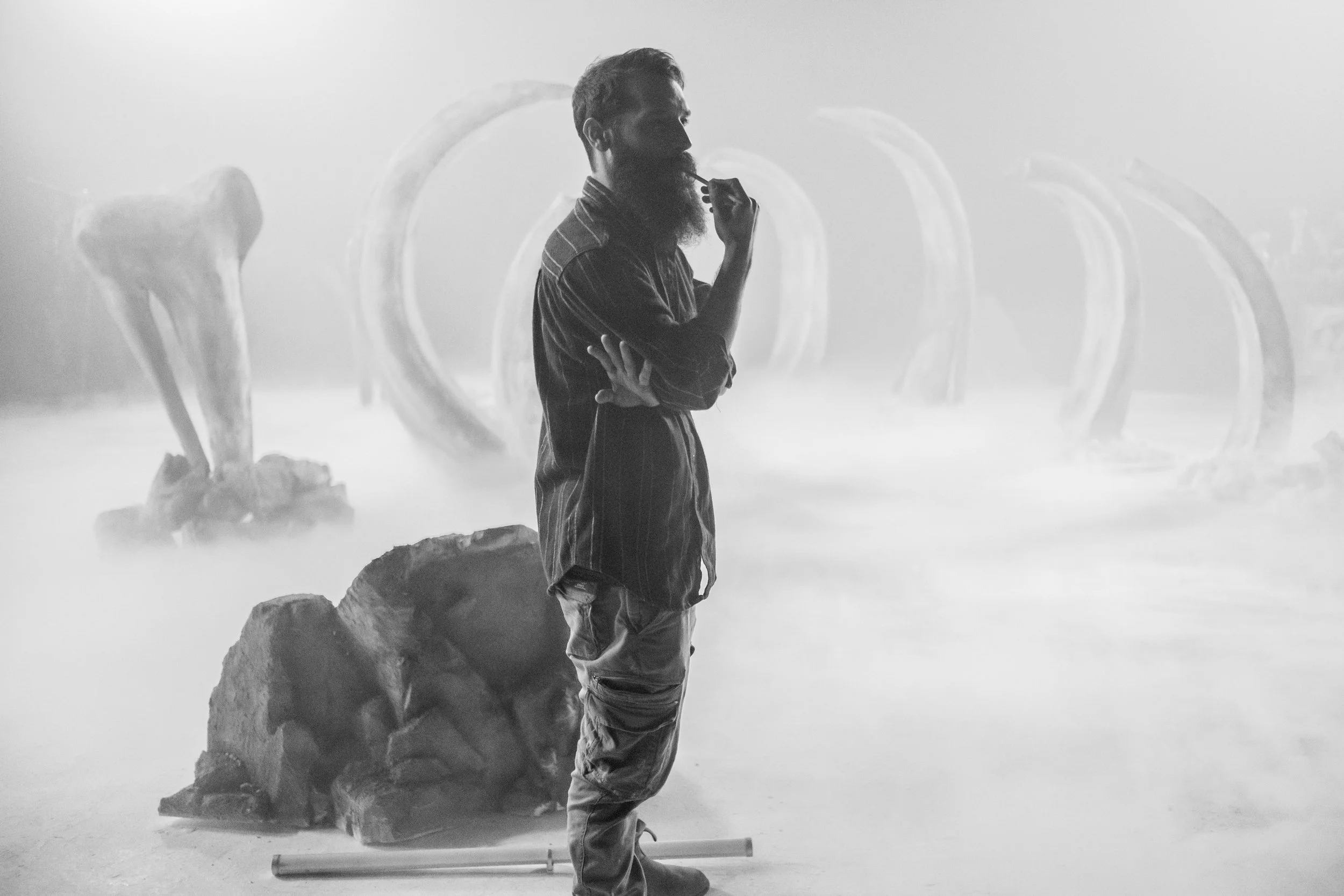 A man with a beard and striped shirt in silhouette, standing with arms crossed and hand on chin, in a foggy setting with art installations or sculptures, including large heart-shaped structures and irregular rocks.music video director , video Clip