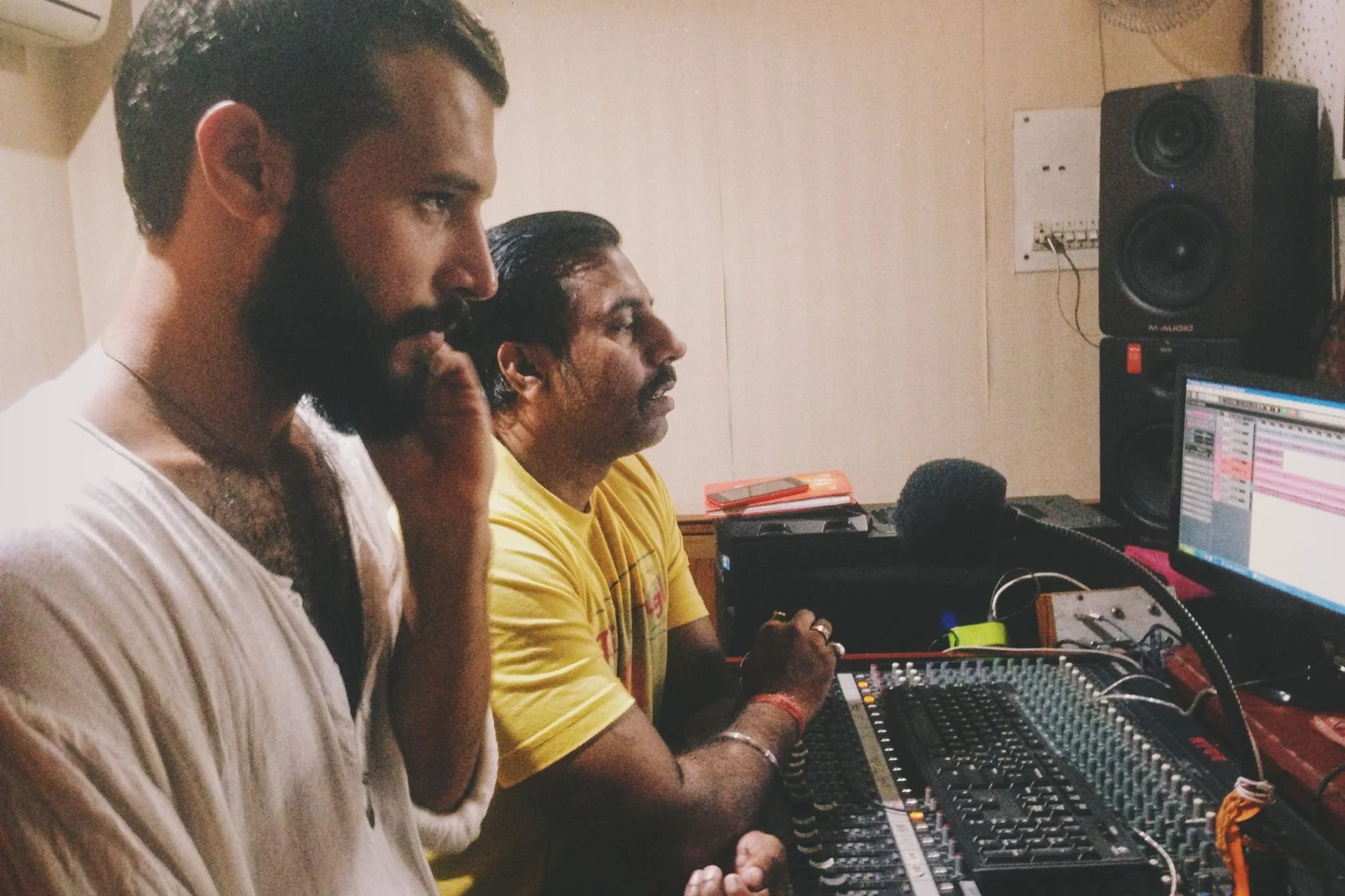 Two men are sitting at a sound mixing console in a recording studio, one is wearing a yellow T-shirt and the other is wearing a striped shirt, both focused on the equipment.