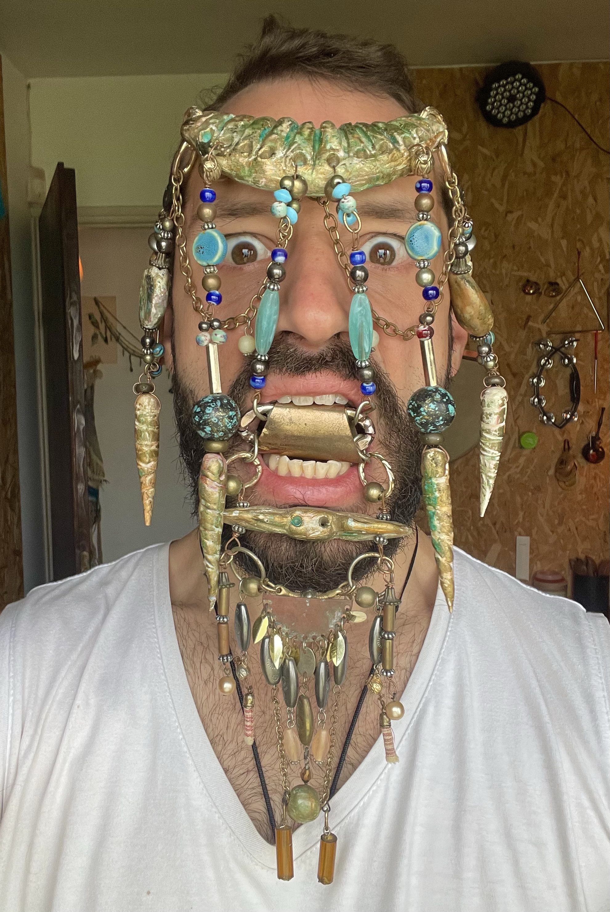 A man with a beard and mustache wearing a white shirt with various necklaces and jewelry covering his face and forehead, including beaded, metallic, and carved items, craft by Avi Mazliah clothes and costumes, Crazy headpiece