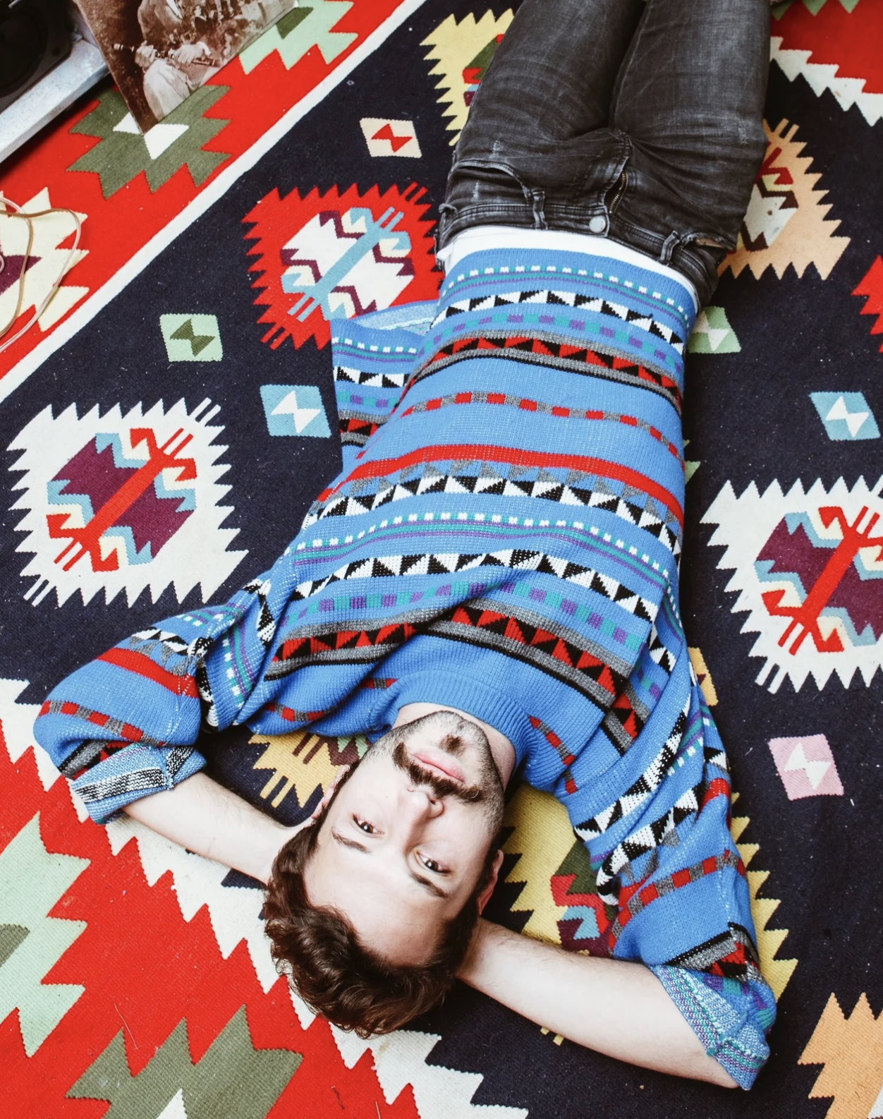 A man lying on a patterned rug with his head resting on his arm and smiling, wearing a colorful patterned sweater and black jeans.