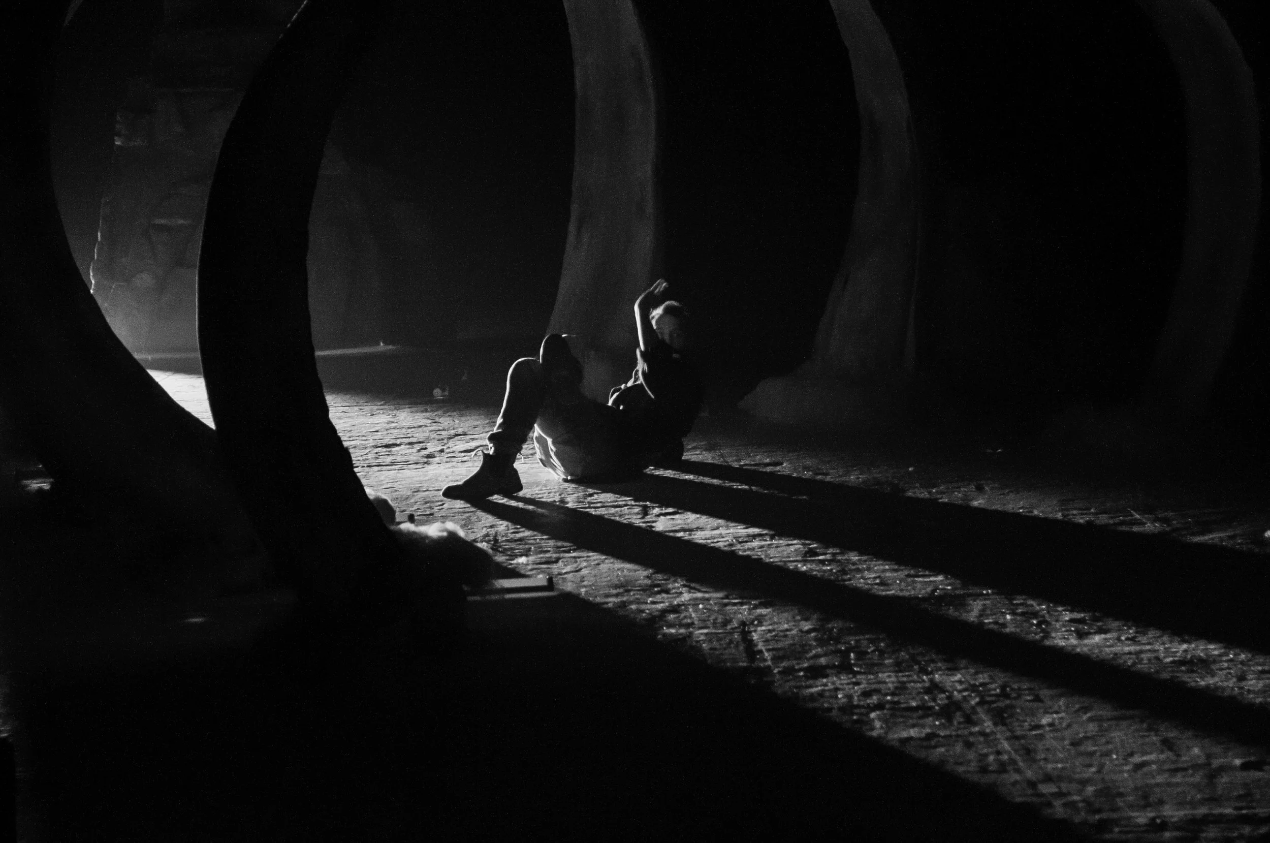 A person sitting on the ground, illuminated by bright light, with large dark structures surrounding them, creating a silhouette effect. music video director , Music video clip