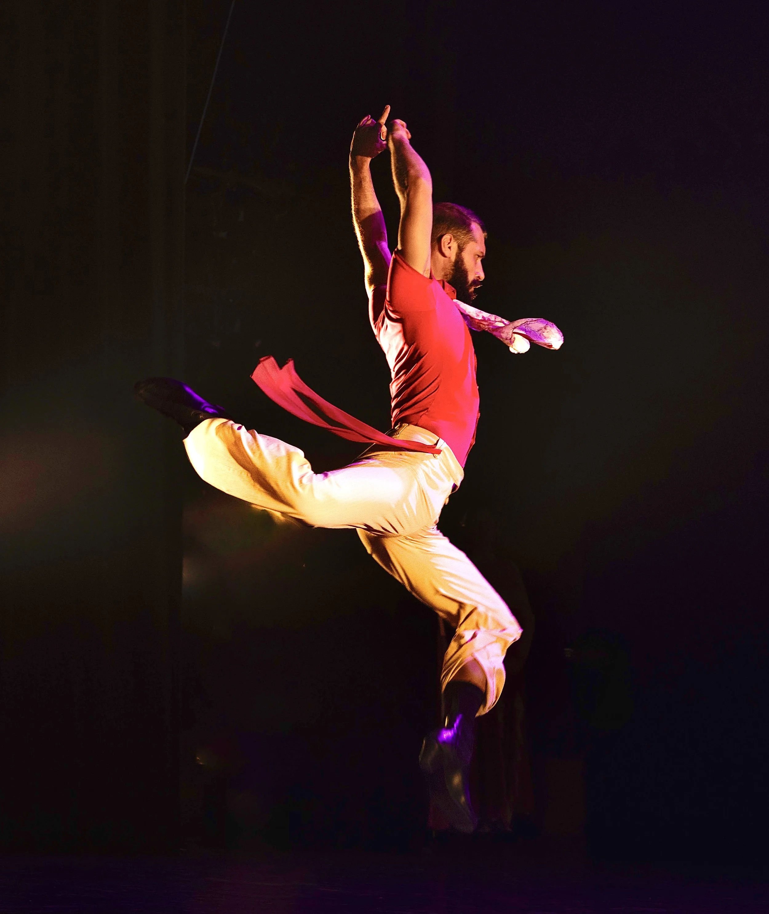 A male performer in a red shirt and beige pants, mid-air in a dynamic dance pose against a dark background, illuminated by purple and pink stage lights. Avi Mazliah dance dancing