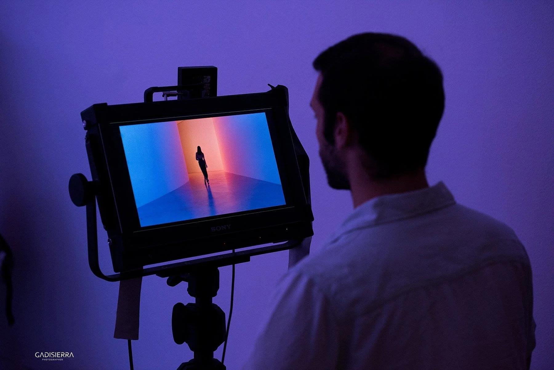 A man watches through a monitor displaying a woman standing in a colorful room with purple, pink, and orange walls. Avi Mazliah on set BTS Omer Adam