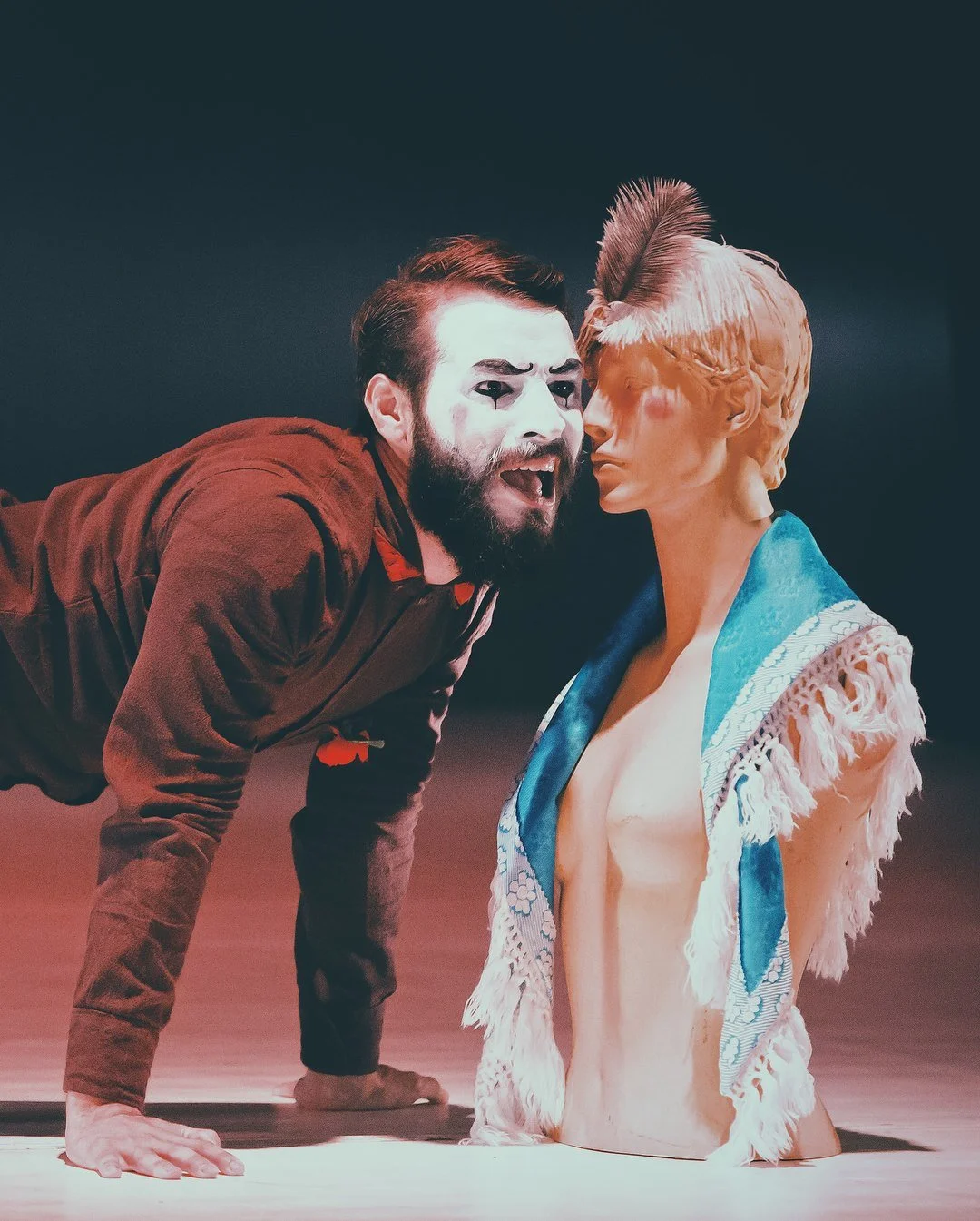 A man with makeup and a beard wearing a red jacket leaning over on one hand, face to face with a mannequin bust of a woman with blonde hair, wearing a blue scarf and a fringed garment, on a stage with a dark background. Avi Mazliah dance dancing