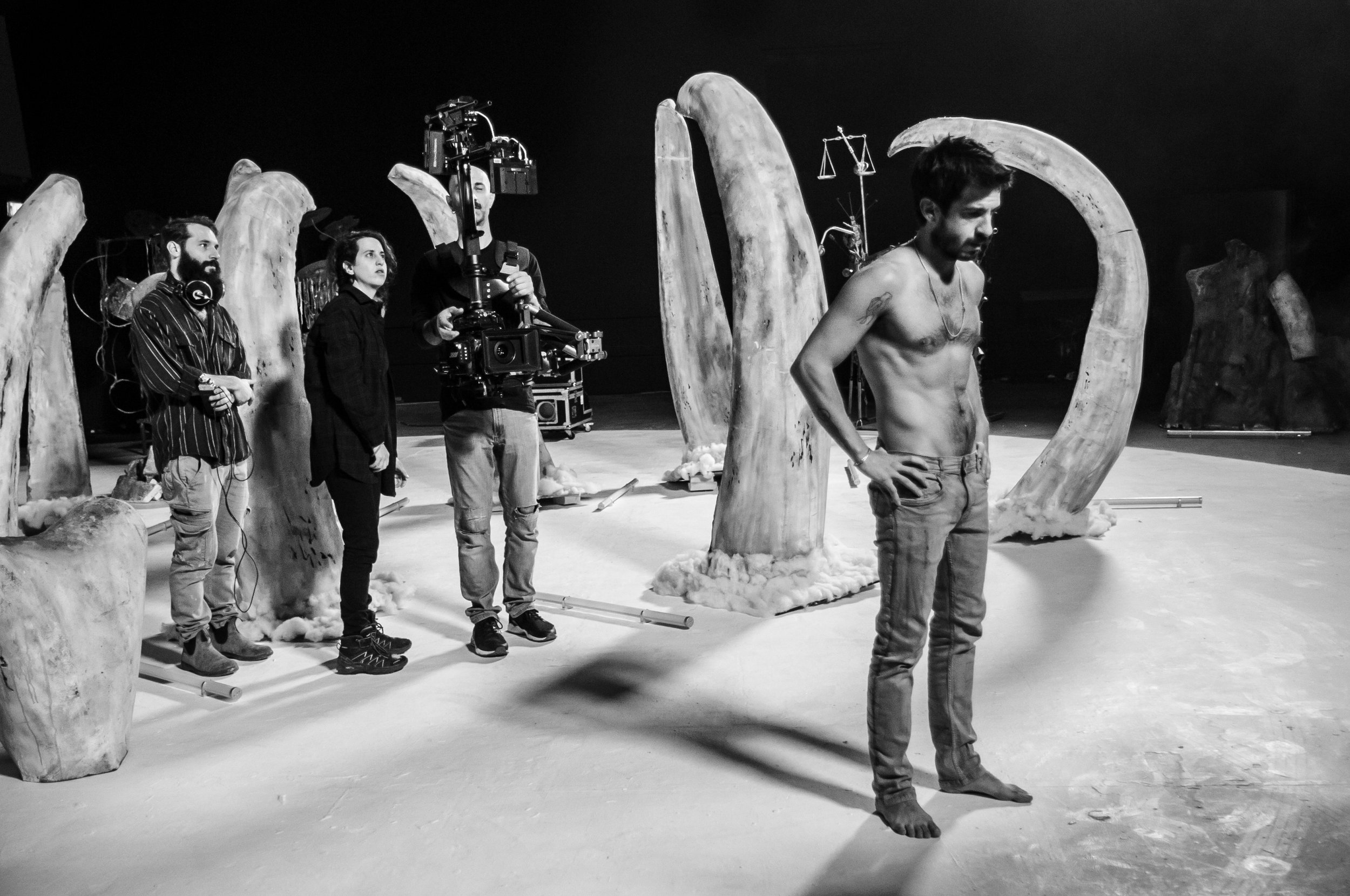 Group of people on a set, with one shirtless man and beautiful art set design and props,  large sculpture elements resembling claws or tusks,
