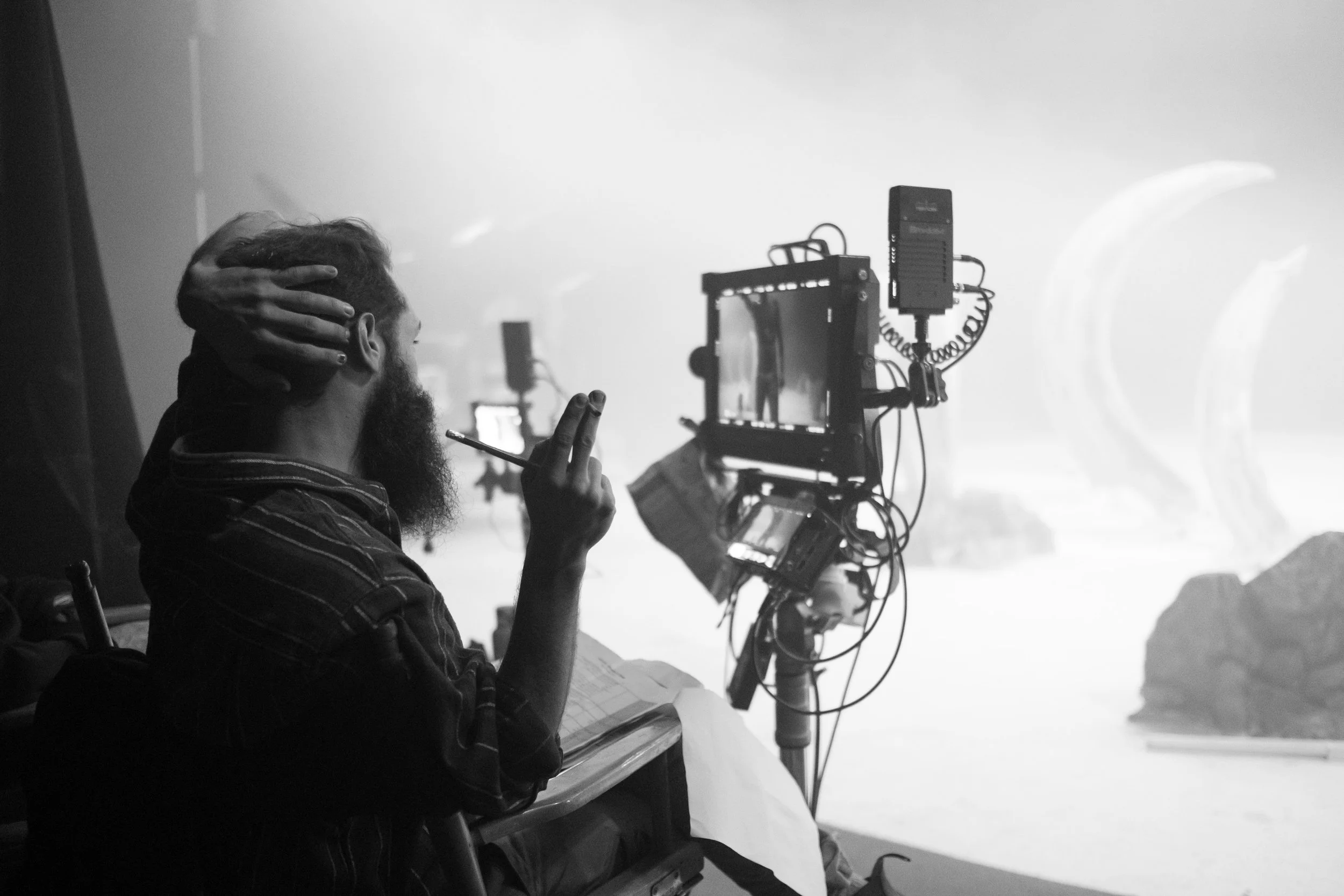 A person with a beard and striped hoodie sitting at a table with a notebook, holding a pen, in front of a film or television camera setup in a studio. The scene is in black and white. mamoota Video clip Avi Mazliah on set BTS