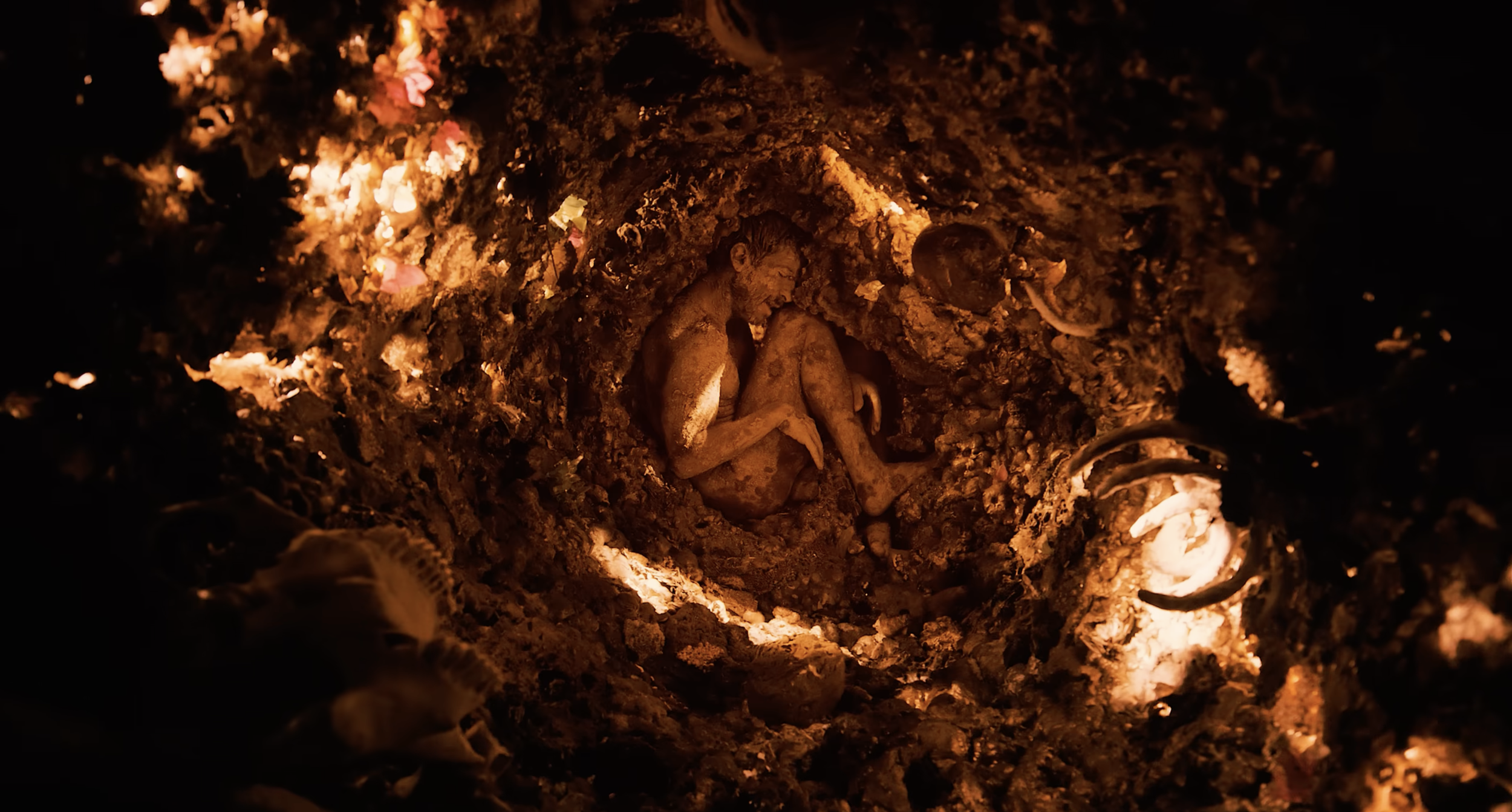 A small, nacked, clay-colored statue of a baby or child wrapped in a cloth, lying in a dirt, surrounded by soil and rocks, inside a dark, underground or cave-like environment. full trunk , mamoota Video clip Avi Mazliah on set BTS