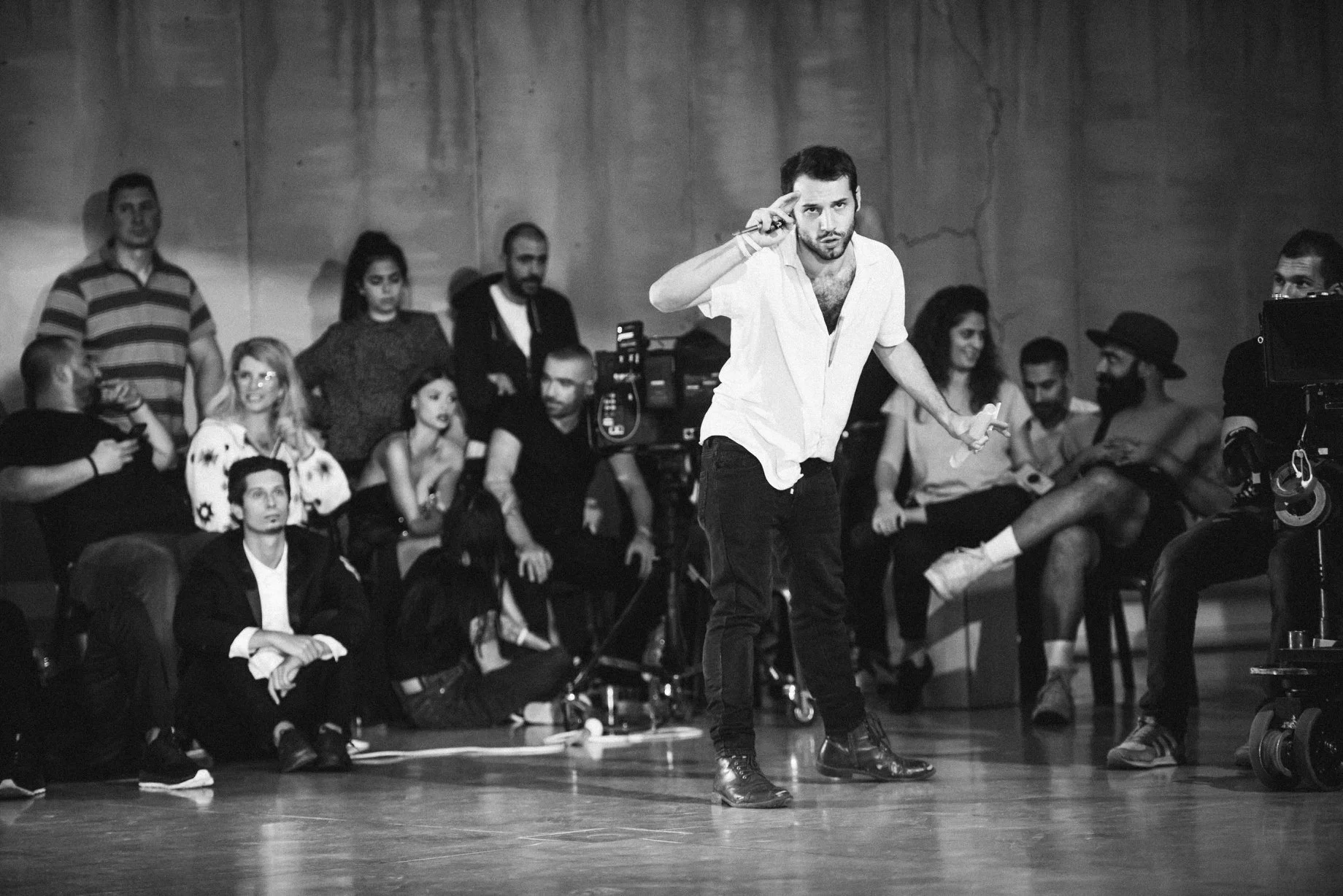 A man in a white shirt and black pants standing on a stage, making a gesture towards his head, surrounded by an audience and film crew in black and white. Avi Mazliah on set BTS Omer Adam 