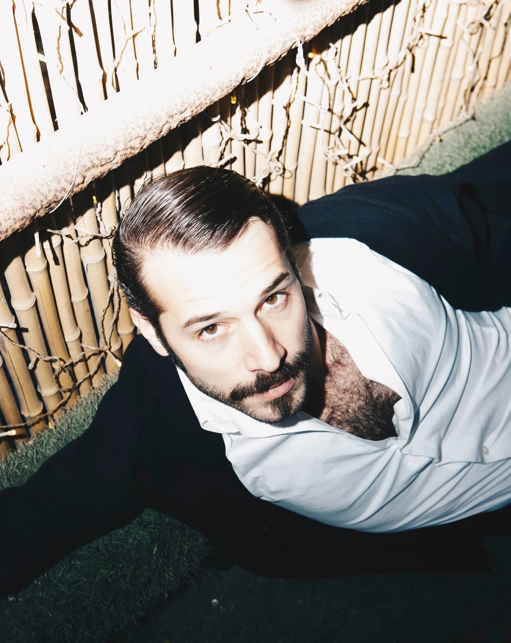 A man with brown hair and a beard lying on the ground with a bamboo backdrop and string lights, wearing a white shirt and a black jacket.